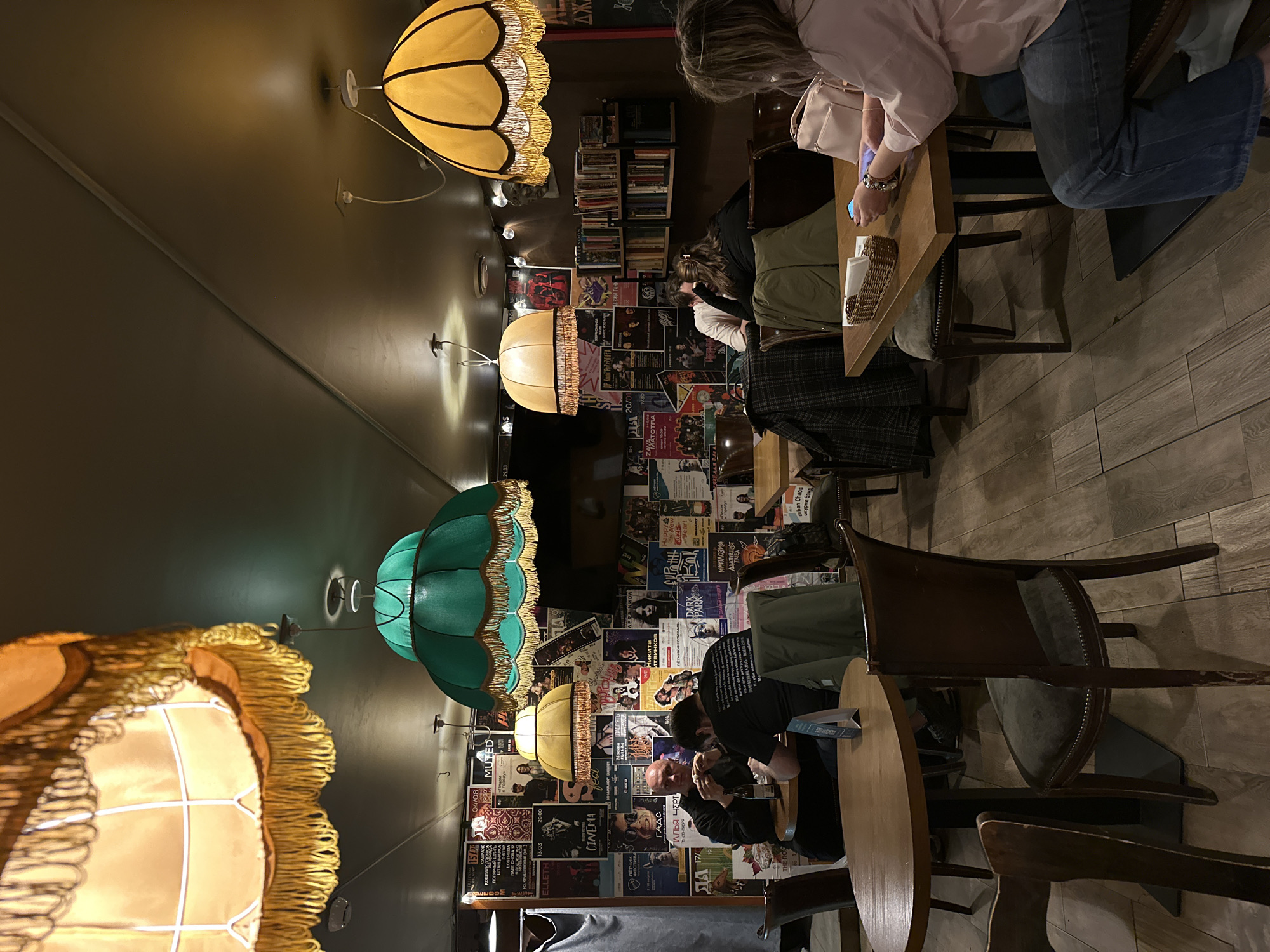 Here is a description of the image:
The image shows the interior of a dimly lit establishment, possibly a café or bar. Several people are seated at wooden tables, some engaging with their phones or each other. The walls are covered in what appears to be a collage of posters, possibly related to music or events. Ornate lampshades in various colors hang from the ceiling, casting a warm glow on the scene. The floor is made of wooden planks. A bookshelf is visible in the background.