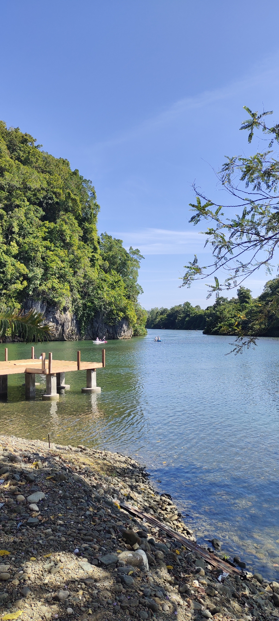 The image depicts a serene river scene, possibly in Lamseunia, Indonesia, given the lush tropical foliage and clear blue sky. In the foreground, a simple wooden pier extends into the tranquil water, supported by concrete pilings. The shore is lined with pebbles and some scattered debris, including a piece of corrugated metal. The river itself is calm and reflects the surrounding greenery. In the middle ground, two small boats with people can be seen navigating the water, suggesting recreational activity. The background is dominated by a steep, tree-covered cliff face on the left, typical of a river gorge. On the right, the river curves into the distance, bordered by dense vegetation. The overall impression is one of natural beauty and peaceful outdoor recreation.