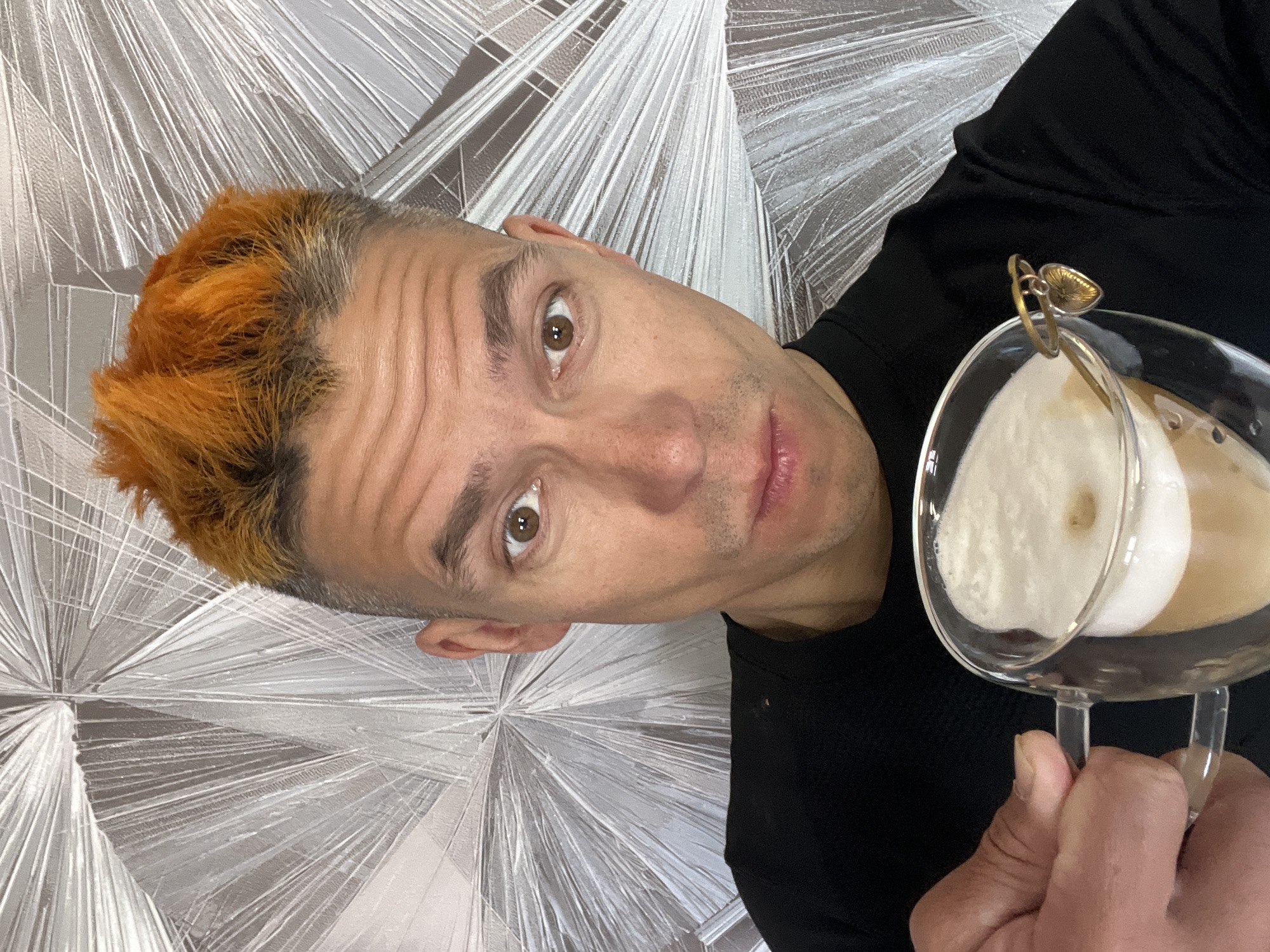 A man with orange hair is holding a glass of foamy coffee in front of a wall with a silver geometric pattern. He is wearing a black shirt. He has brown eyes and a light skin tone. The location is likely indoors. The image does not provide enough information to confirm the location in Chaikovsky, Russia.