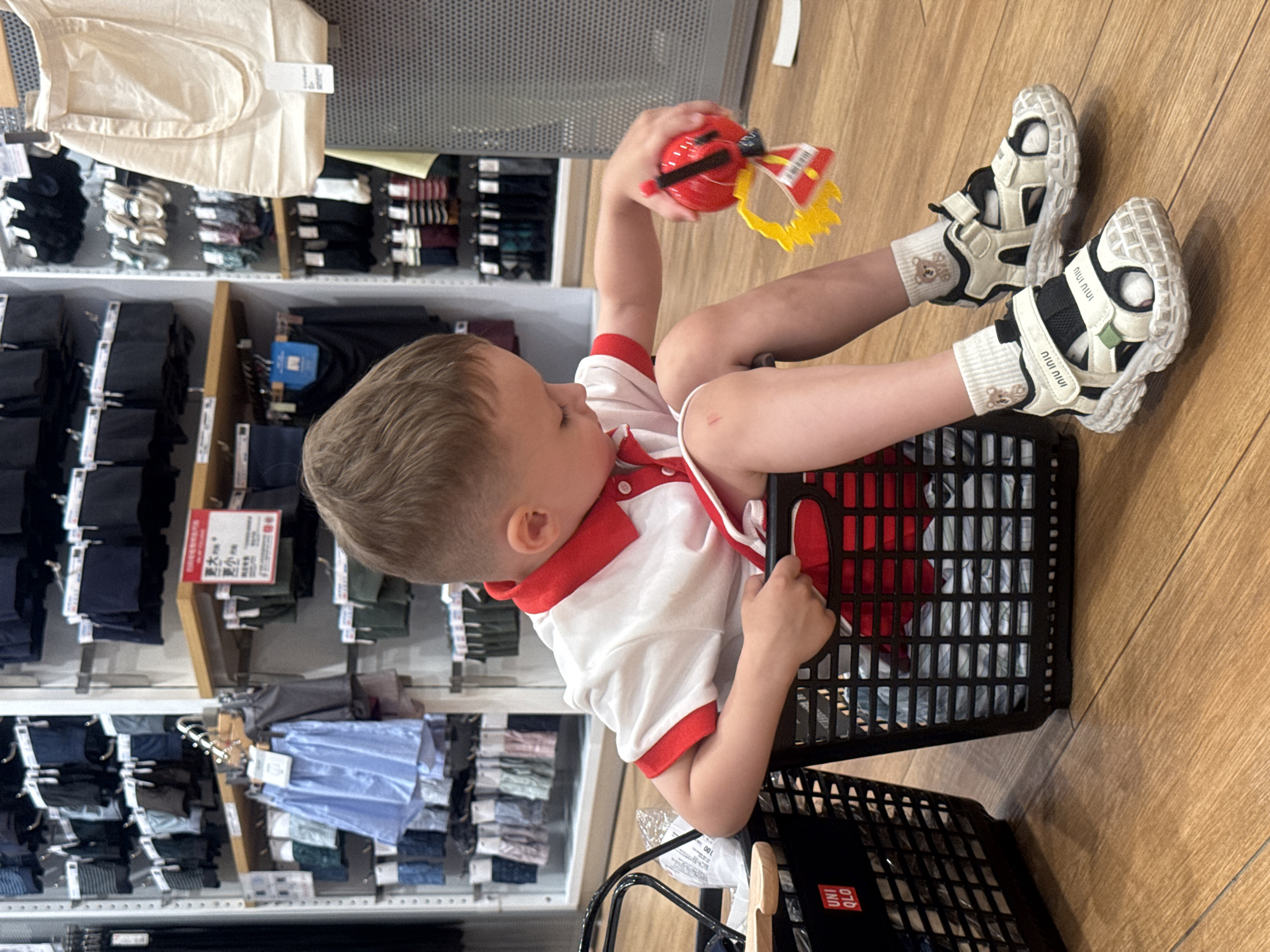 A young Caucasian boy with blonde hair sits in a black plastic shopping basket in a retail store. He is wearing a white polo shirt with red trim, grey shorts, white socks with a teddy bear pattern, and white and black sandals. The boy is holding a red and yellow toy in his right hand. The basket is filled with folded clothes, some of which appear to be plaid. Another black shopping basket is visible to the left. The background shows shelves stocked with clothing, primarily underwear and socks, in various colors. A light-colored canvas tote bag is hanging from a shelf on the right. The flooring is a light brown wood. The store appears to be a UNIQLO, indicated by the logo on the left basket. The location is a UNIQLO store in Qinhuangdao, China.
