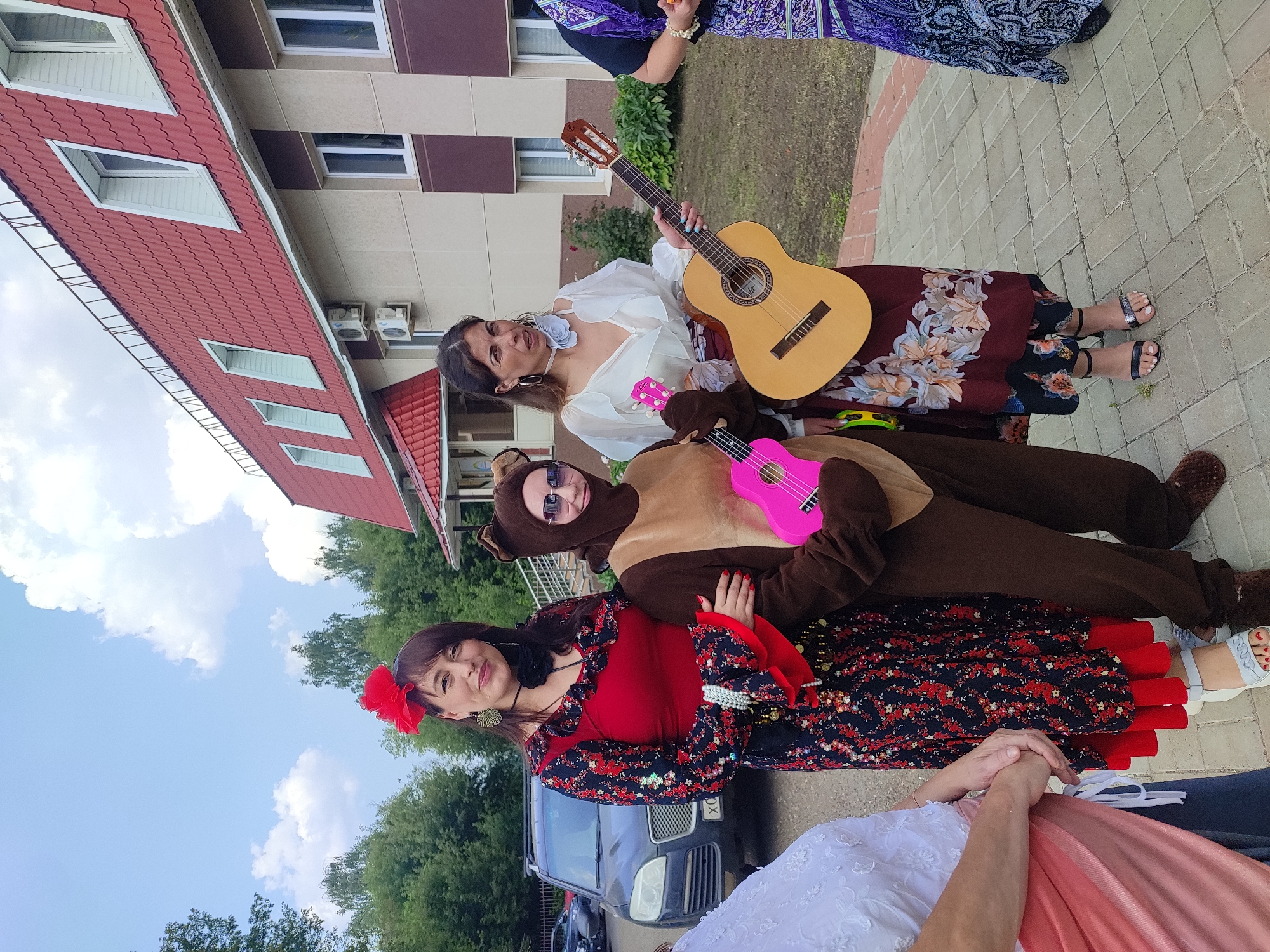 Three individuals are posing for a photo outdoors in front of a building with a red, tiled roof. The person on the left, dressed in a traditional red and black floral pattern costume with a red flower in their hair, stands next to a person in a brown bear costume, who is holding a pink ukulele. The third person on the right is wearing a white blouse and a long, floral skirt, and is holding an acoustic guitar. The building appears to be a commercial or administrative building, possibly in a suburban or semi-rural area. The background includes trees and a parked car, suggesting an outdoor event or gathering. The location is identified as Naberezhnye Chelny, Russia.