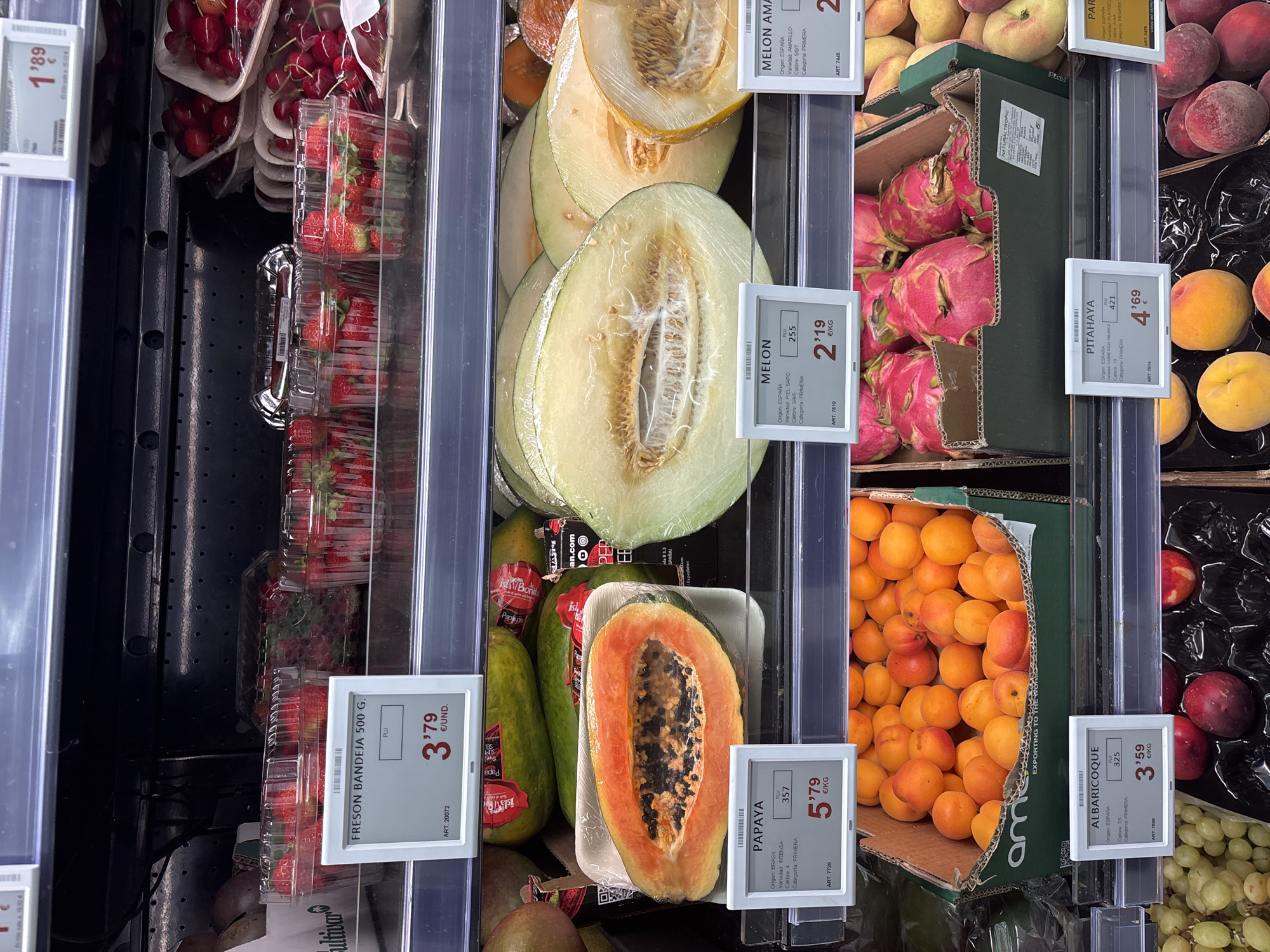 This is an eye-level, medium shot of a supermarket produce aisle in Urbanización Villa Martín, Spain. The image showcases various fruits displayed on refrigerated shelves. From top to bottom, the shelves contain: strawberries in plastic containers, ripe cherries in red-lined containers, and a large papaya cut in half, revealing its seeds. Below the papaya are several halved cantaloupes, and to their right, a halved yellow melon. Further down, a cardboard box is filled with ripe apricots. The bottom shelf displays red dragon fruits and peaches. Each fruit display has a digital price tag indicating the item name, origin, variety, and price per kilogram or unit. The lighting is bright, typical of a supermarket, and the shelves are clean and well-stocked. There are no people visible in the frame.