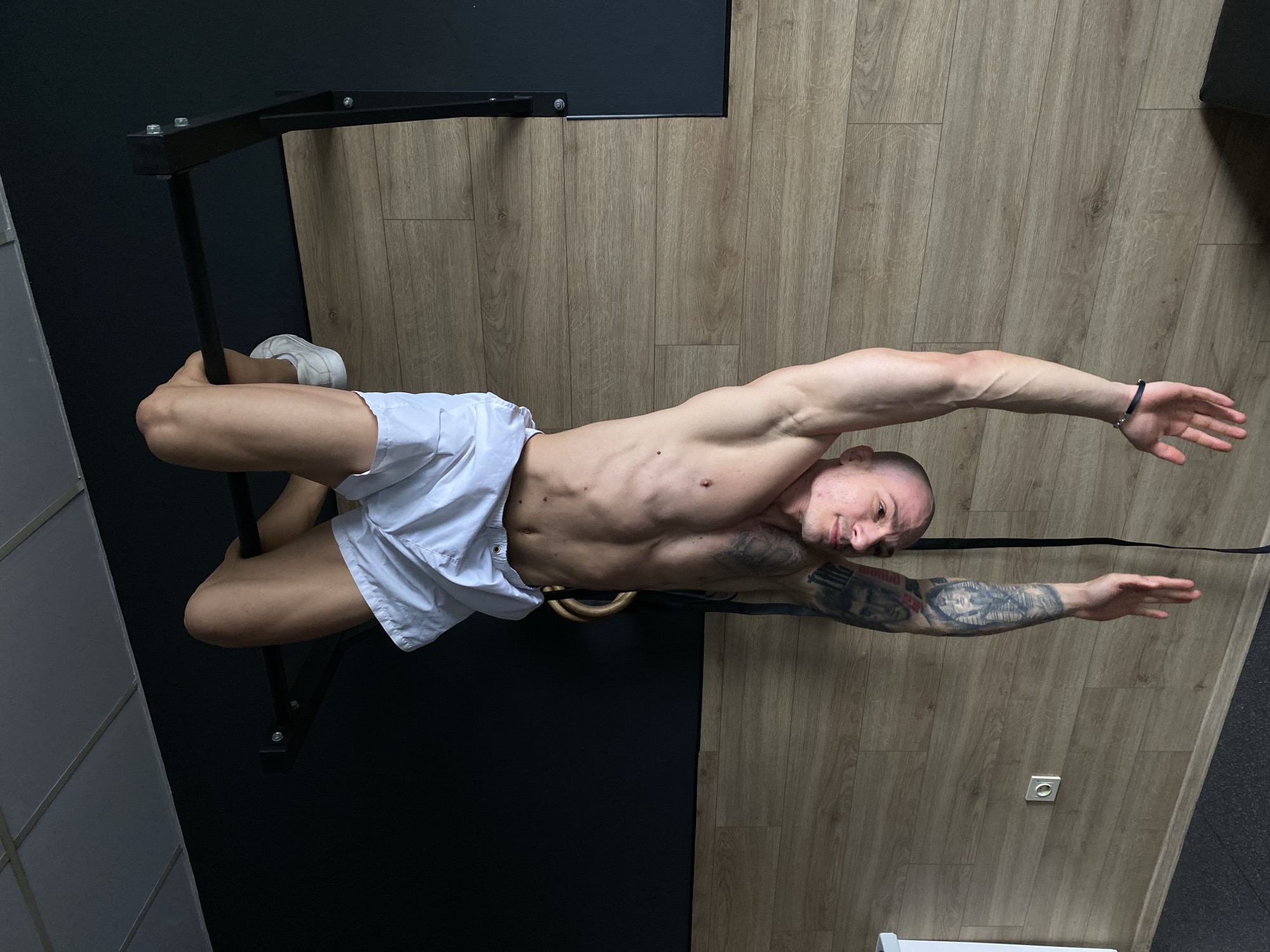 A male individual is depicted suspended upside down by his knees from a black horizontal pull-up bar in a gym located in Pleven, Bulgaria. The subject has a shaved head, light skin, and extensive tattoos on his left arm, including a portrait and an Egyptian pharaoh. He is shirtless, revealing a toned physique, and wears white shorts and white sneakers. A thin black bracelet is visible on his right wrist. His arms are extended downwards with open palms, and he looks directly at the viewer. The background wall features a dark blue/grey upper section and a lower section clad in light brown wood laminate, which also covers the floor. A white grid-style drop ceiling is overhead. A metallic power outlet is present on the wooden wall section to the left. Gymnastic rings with black straps and an additional black strap hang from the pull-up bar behind the individual. The floor transitions to a darker, possibly rubberized, material in the bottom left.