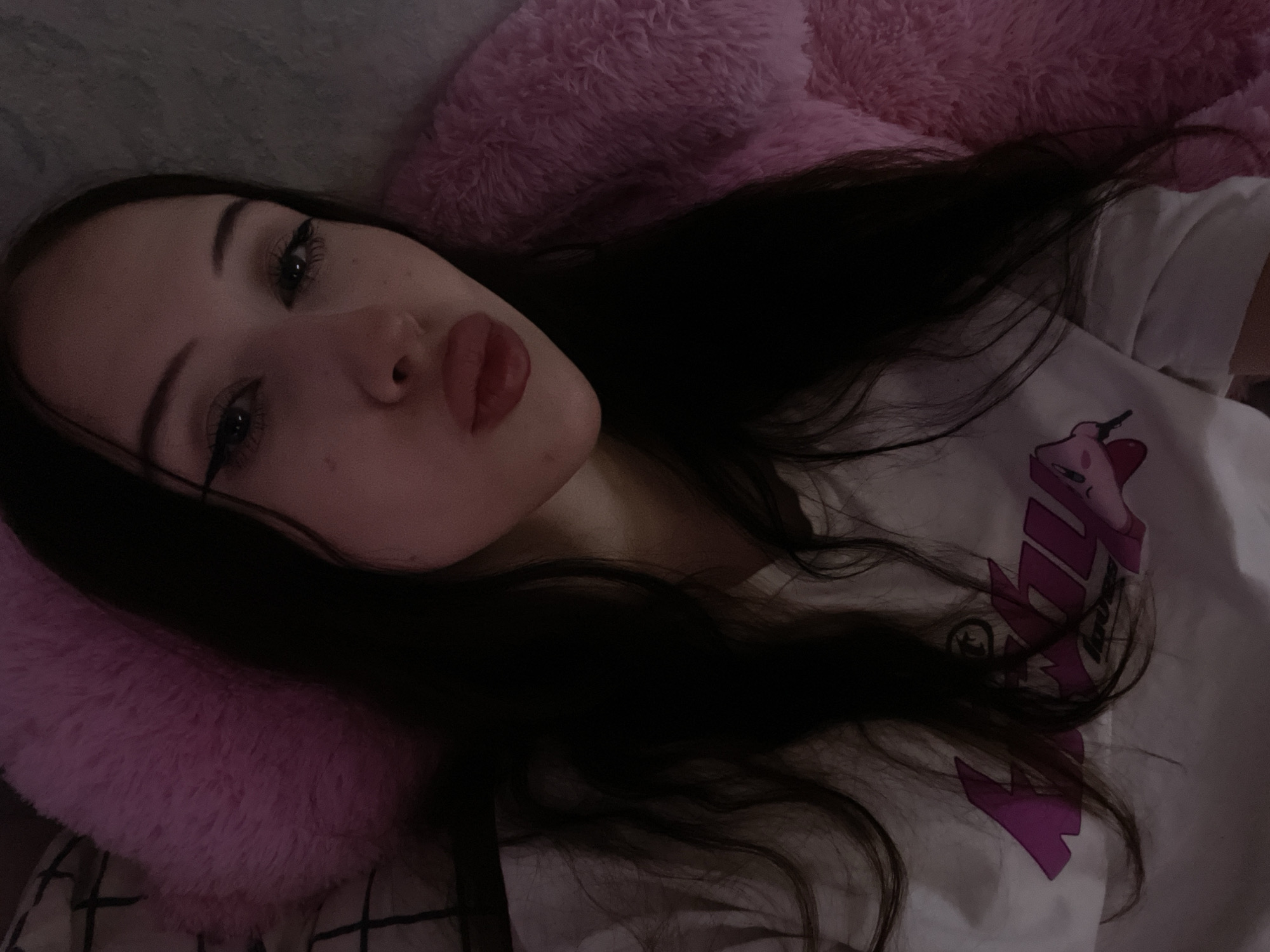 A young woman with long, dark hair is lying on her side, propped up by a fluffy pink pillow. She is wearing a white t-shirt with a pink graphic design. Her head is tilted back slightly, and her gaze is directed towards the camera. She is wearing makeup, including winged eyeliner and lipstick. Her expression appears relaxed or playful. The lighting is dim, suggesting it might be nighttime or a dimly lit room. The background is composed of what appears to be bedding. There is no information about the specific location beyond it being Votkinsk, Russia.