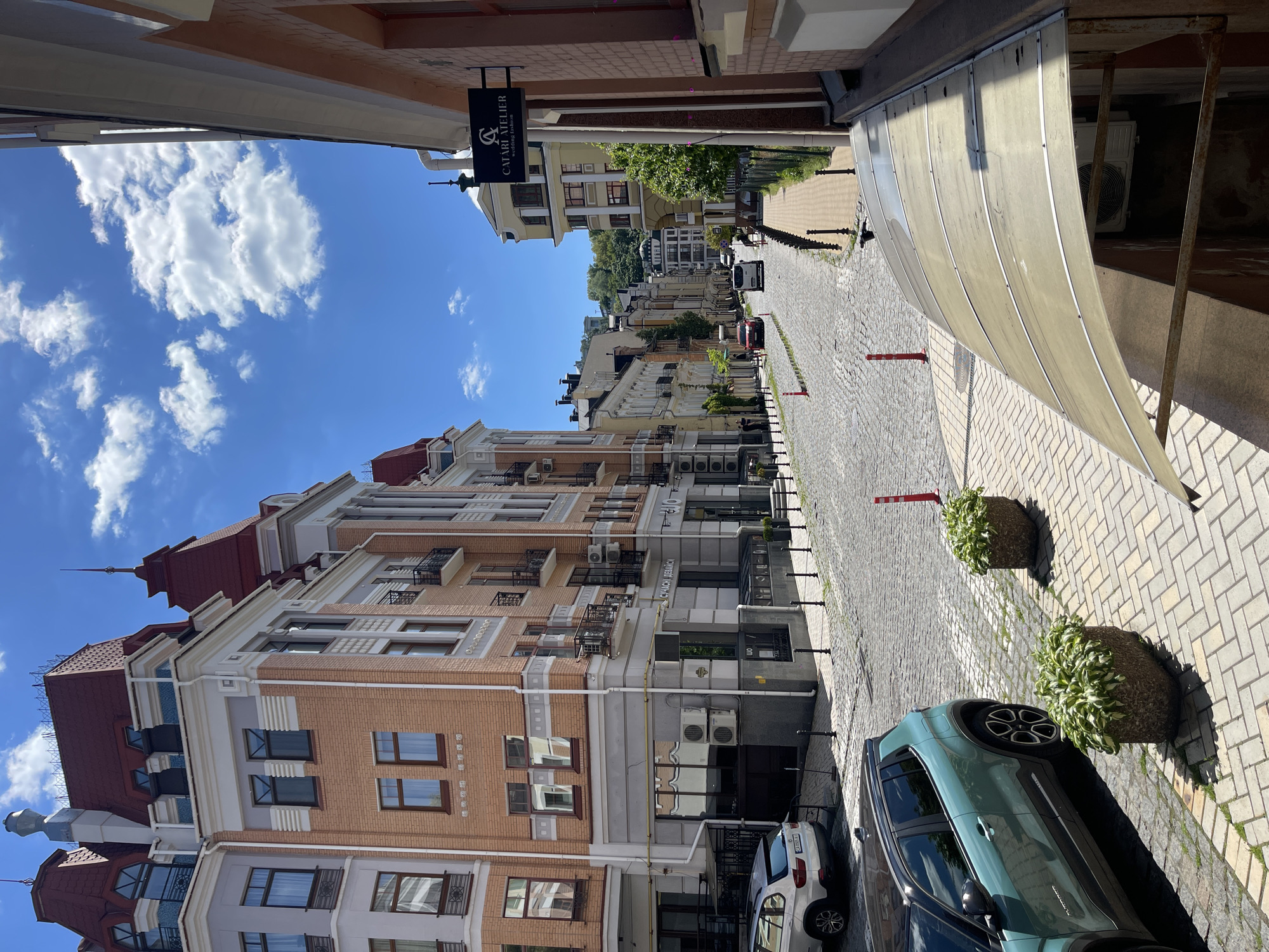 The image displays a sunlit street scene in Kyiv, Ukraine, looking down a gently sloping street. The time of day appears to be mid-morning to early afternoon, characterized by a bright blue sky with scattered white cumulus clouds and clear shadows.
The street itself is paved with cobblestones, bordered by a lighter-colored tiled sidewalk. Multi-story buildings line both sides of the street, featuring traditional European architecture. The buildings on the left have light brown brick facades, white trim, decorative elements, and distinctive reddish-brown tiled roofs, some with spires. Numerous air conditioning units are visible on their exteriors.
Along the right side of the street, a large, curved, translucent awning extends over part of the sidewalk. Planters filled with lush green foliage are spaced along the sidewalk, alongside red and white bollards. Several cars are parked along the street, including a prominent dark teal/green SUV in the foreground on the right, and a white car behind it. Other vehicles are visible further down the street as it recedes into the distance, revealing more buildings and green hills.
A black hanging sign is visible on the left-hand building, displaying the text "CATART ATELIER" and "wedding dresses," along with a stylized 'C' and 'A' logo. No people are visible in the scene, contributing to a tranquil urban atmosphere.