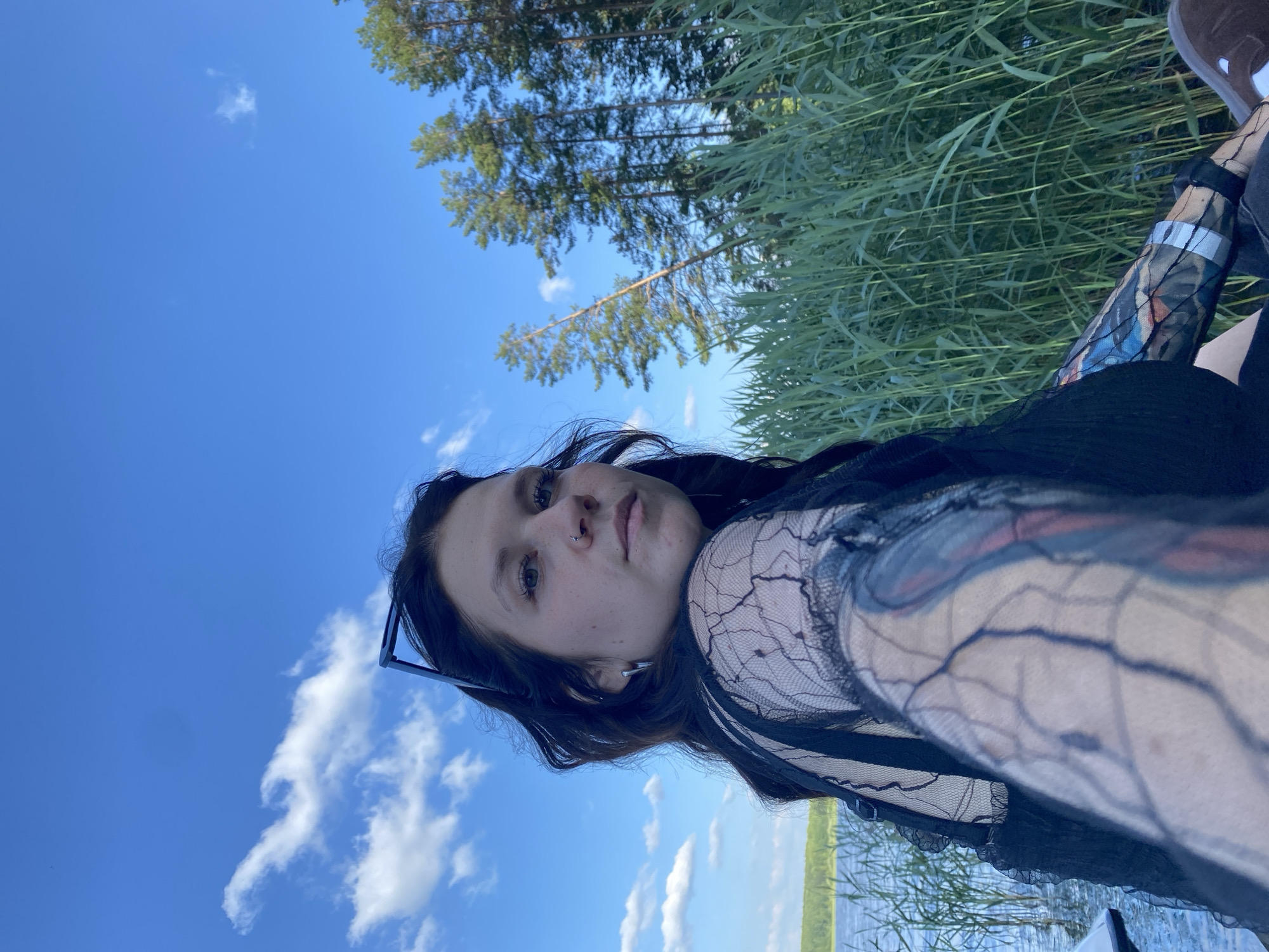 A person is shown from the chest up, in a close-up shot, outdoors, with a lake and trees in the background. The person has dark hair, a nose ring, and visible tattoos on their arm. They are wearing a black lace top. The sky is blue with some white clouds, and the sun appears to be shining. The location is the city of Chaykovsky, Russia.