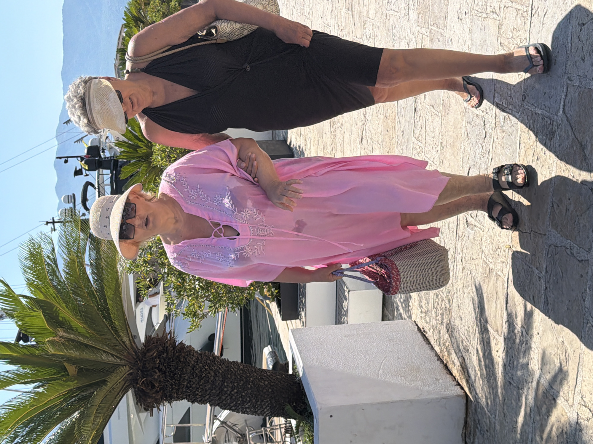 Two women walk along a paved walkway near a body of water. The woman on the left wears a pink, loose-fitting cover-up with white embroidery and a wide-brimmed hat. She carries a woven tote bag. The woman on the right wears a black, sleeveless dress and a visor. They are walking arm-in-arm. Behind them, a large yacht is docked, and palm trees and lush greenery are visible. The background features a mountainous landscape under a clear blue sky. The walkway appears to be made of stone pavers with visible grout lines. The scene suggests a sunny day in a waterfront setting, likely a marina or promenade in Budva, Montenegro.