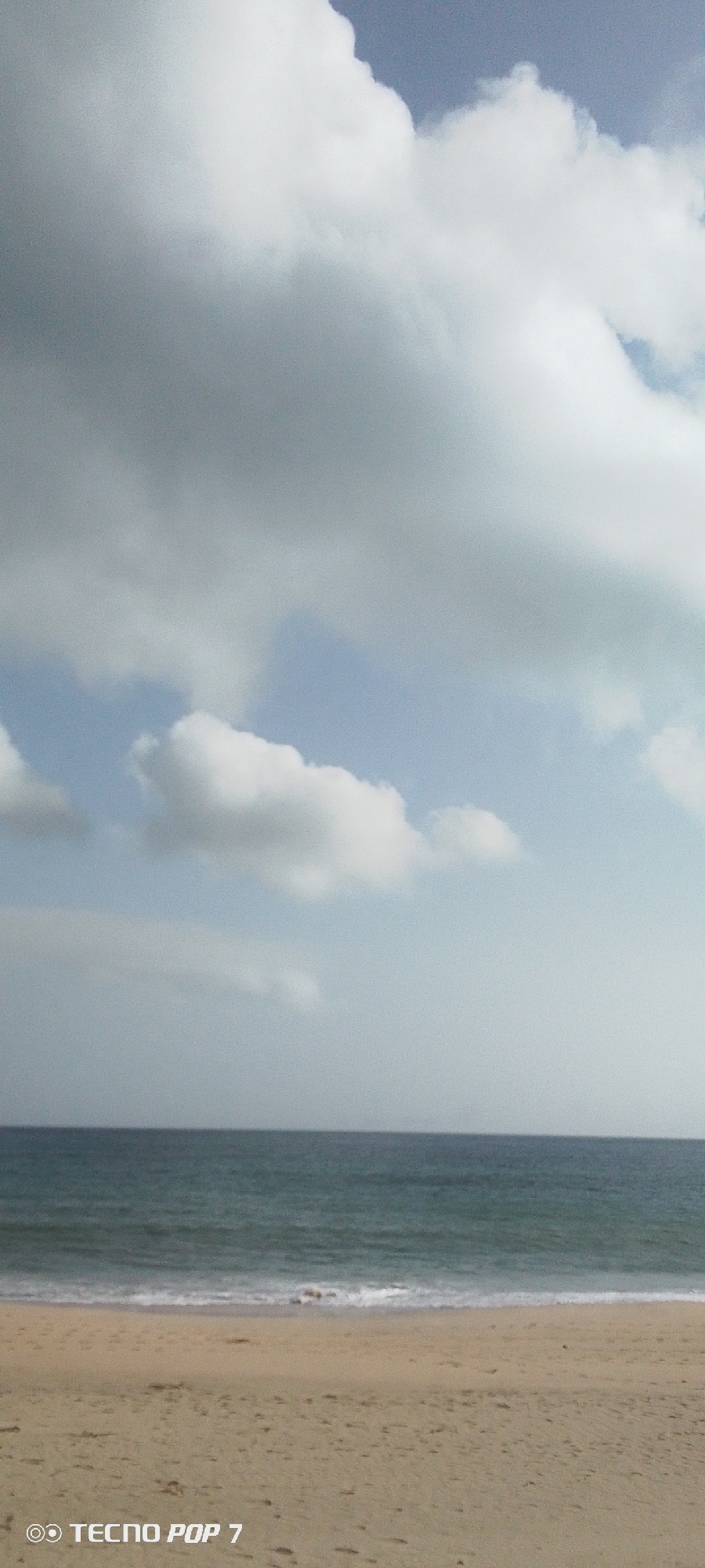 The image shows a beach scene, likely in Pedro González, Venezuela, though specific location details are not discernible. In the foreground is a sandy beach with a gentle slope. In the midground, the ocean meets the shore with small waves visible. The background is filled with a sky dominated by clouds, with some patches of blue sky.