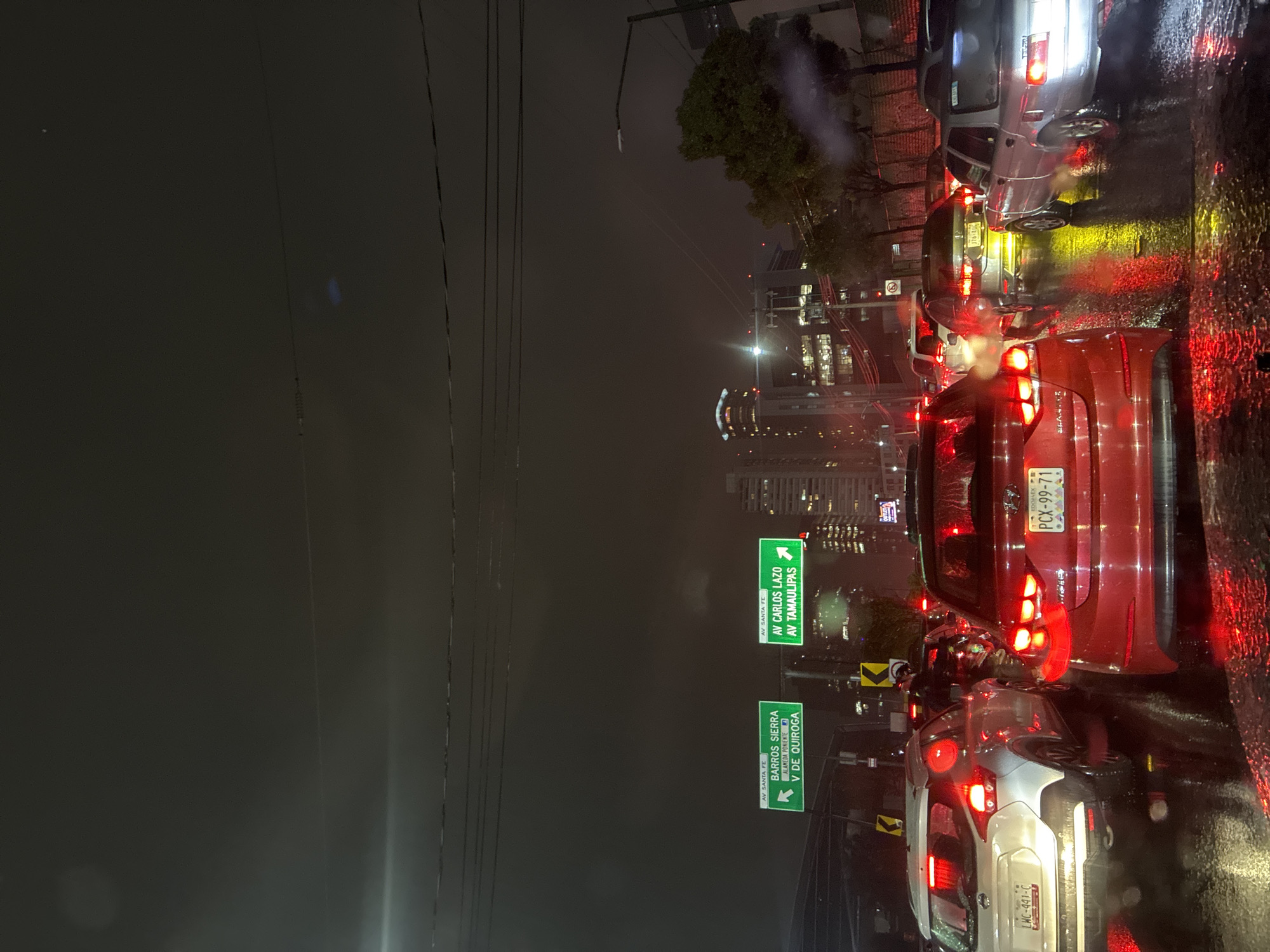 Here is a description of the image:
The image shows a congested city street at night, likely during or after a rainstorm, based on the wet pavement and reflections. It appears to be taken in Mexico City, based on the license plate, which features the word "EDOMEX". The congestion is focused in one direction. There are street signs that denote direction to the "Barros Sierra Alameda Poniente y de Quiroga" and to "Av Carlos Lazo Av Tamaulipas". The traffic consists of passenger vehicles, and the vehicles are illuminated by their tail lights.