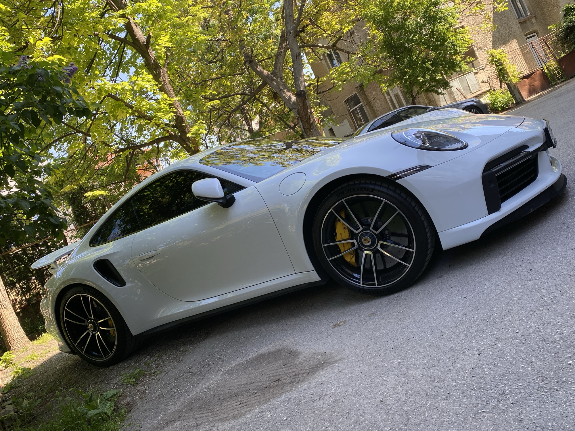 A white Porsche 911 Turbo S is parked on an asphalt road on a sunny day. The car has black rims, yellow brake calipers, and a black roof. Trees with green leaves are in the background, partially obscuring a building with windows and a rusty gate. Another dark-colored car is partially visible behind the white Porsche. The road surface shows signs of wear and contains a dark wet spot. The location is inferred to be Pleven, Bulgaria, based on external information not present in the image.