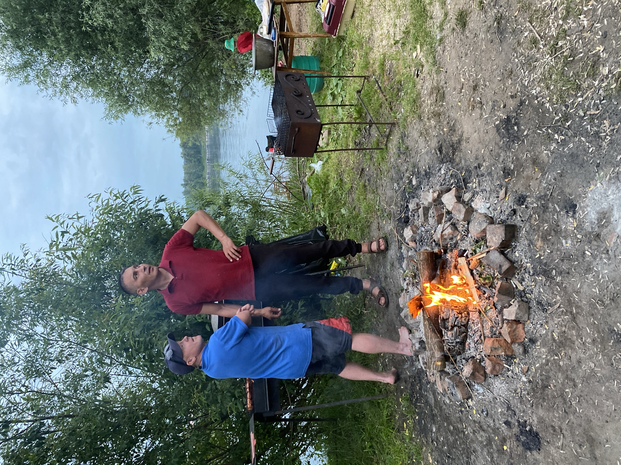 Two men are by a campfire and a grill near a body of water in Noviy, Russia. The man on the left wears a blue polo shirt and red and black shorts, and is barefoot. The man on the right wears a red polo shirt and black pants. He is wearing sandals. A fire is burning in a makeshift pit of bricks. A grill with food is to the left of the men. To the right, a table holds a large pot and other items. Trees and water are in the background.