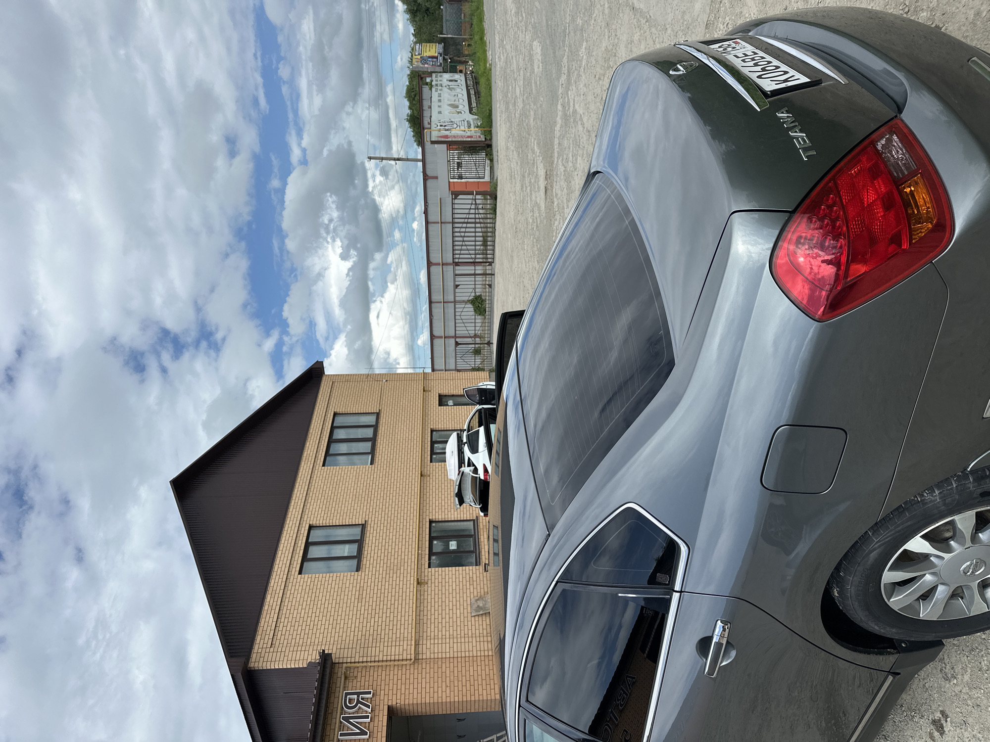 The image captures a grey Nissan Teana sedan parked outdoors in Orsk, Russia. The car is viewed from the rear-right quarter. In the background, a brick building with a dark-colored metal roof is visible, with several windows and an entrance. A white car with a roof box is parked in front of the building. Further in the distance, there is a fenced area with a metal structure and some advertising posters on a wall. The sky is partly cloudy with patches of blue. The ground appears to be a dusty or gravelly surface. The license plate of the Nissan Teana is partially visible and reads "K066BE 156".