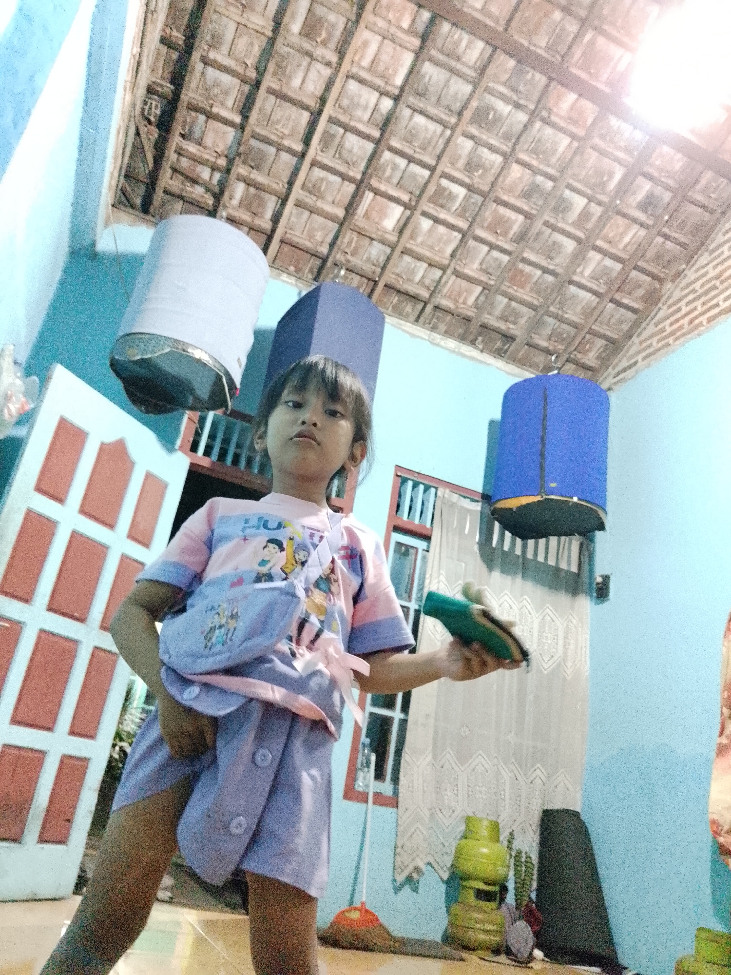 In an indoor scene with light blue walls and a wooden plank ceiling, a young girl stands. She is wearing a pink and blue t-shirt with cartoon characters and a matching purple skirt with buttons. A light purple fanny pack is slung across her chest, tied with a pink ribbon. In her right hand, she holds a green object, possibly a toy or a tool. To her left, a white door with red accents is partially visible. Above, two cylindrical objects hang from the ceiling: one is white, and the other is blue. In the background, a patterned white curtain hangs behind a green gas cylinder and a black conical object. The overall impression is of a domestic setting, likely a home, in Kecamatan Pati, Indonesia.