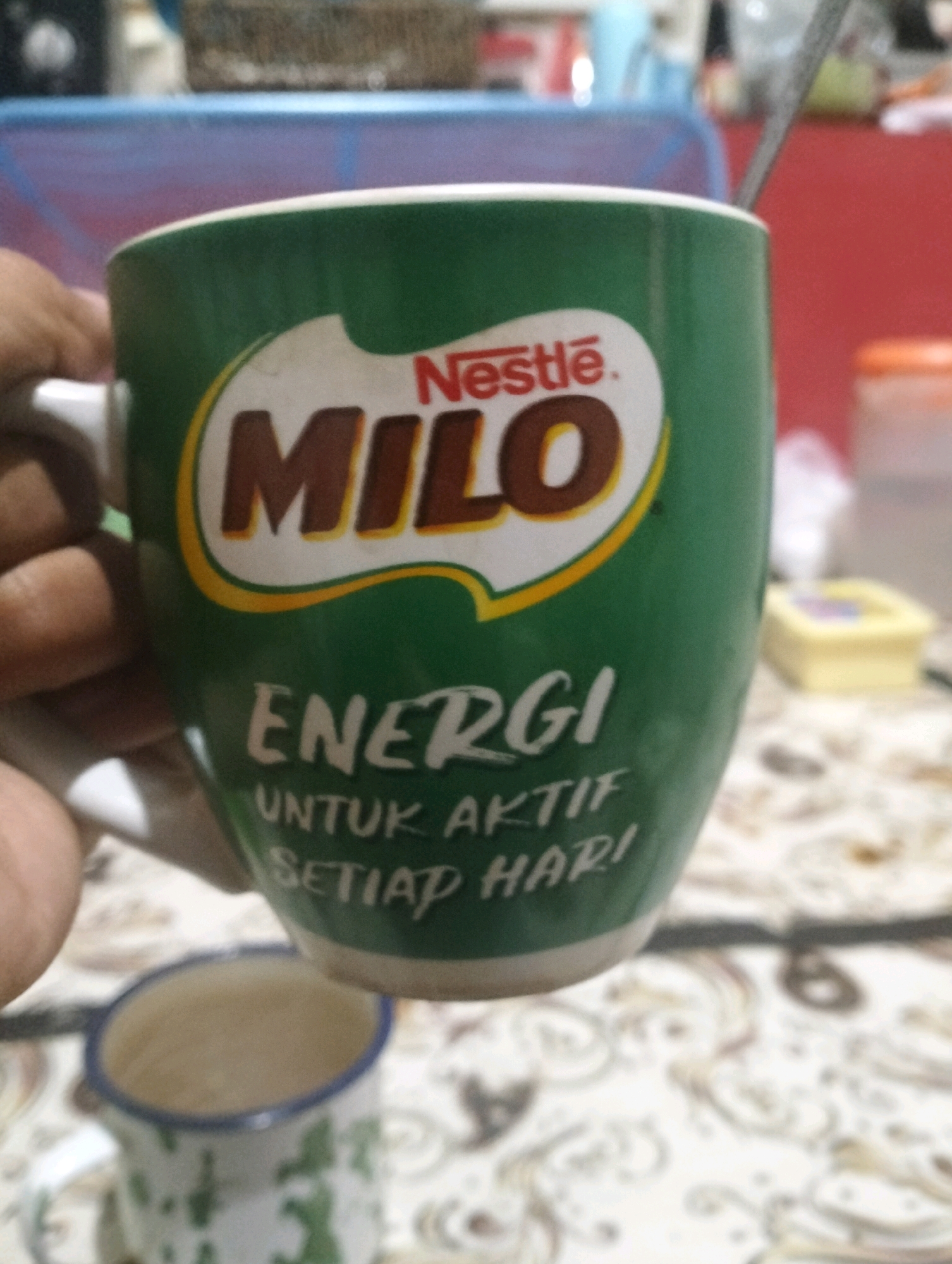 A green Milo mug is held by a person's left hand. The mug has the Milo logo with "Nestlé" above and "Milo" written in large brown letters. Below the logo, the words "ENERGI UNTUK AKTIF SETIAP HARI" are written in white text. A spoon is visible in the mug. In the background, a blurry scene shows a red wall and some objects on a patterned surface. A smaller, white mug with green patterns is also visible in the foreground. The image is a close-up shot of the Milo mug, with the background out of focus. The location is not specified in the image.