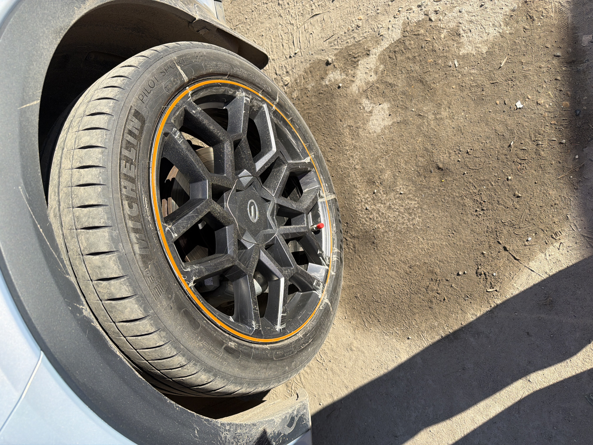 The image displays a close-up, high-angle view of a dirty car wheel and tire, situated on dusty ground under bright sunlight, casting distinct shadows. The scene is set outdoors, in Chaikovsky, Russia.
The primary subject is the vehicle's front wheel, which features a dark, multi-spoke alloy rim. A notable detail is the orange decorative strip encircling the rim, which shows visible scuffs and scratches. The rim itself is covered in dust and appears to have numerous superficial marks. A red valve stem cap is present on the rim.
The tire is clearly identifiable as a Michelin Pilot Sport 4. "MICHELIN" and "PILOT SPORT 4" are prominently visible on the sidewall, along with partial text like "OUTSIDE" and "TEMPERATURE". The tire's tread pattern is visible and appears moderately worn, also coated in a layer of dust.
The wheel is positioned beneath the car's fender, which includes a light-colored body panel (likely silver or light gray) and a contrasting dark, possibly black, plastic fender flare. Both vehicle components are noticeably dirty, with streaks and accumulations of dust and grime, indicating the car has been driven in dusty conditions.
The ground surface visible in the image is dry and uneven, composed of dirt, small stones, and possibly some patches of harder earth or worn concrete. The overall impression is of a vehicle in a somewhat rugged or unpaved outdoor environment.
