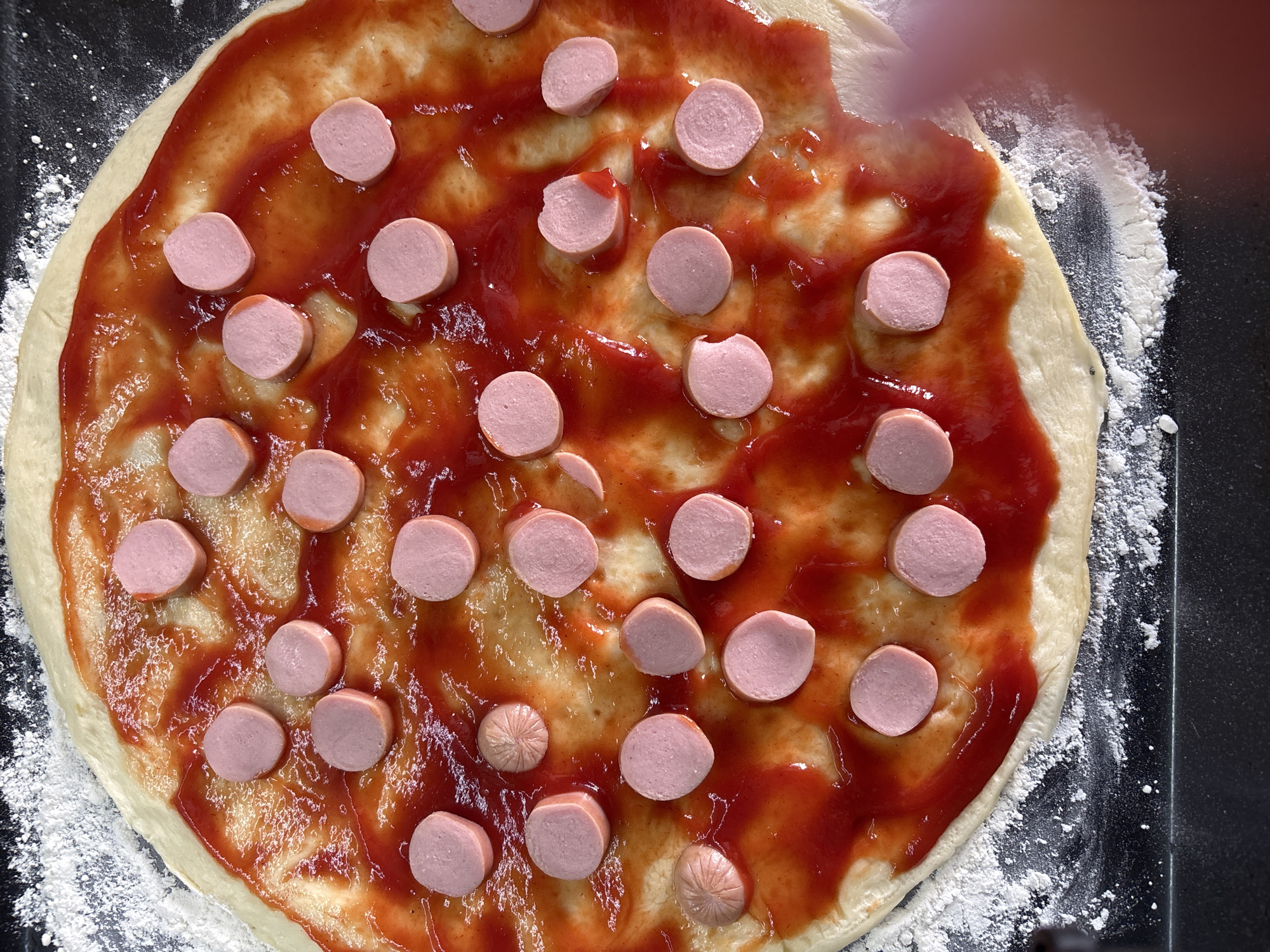 This is an overhead shot of a pizza being prepared in Bila Tserkva, Ukraine. The pizza dough is spread thinly on a dark surface, possibly a baking sheet, dusted with flour. A generous layer of red sauce, likely tomato sauce, covers most of the dough. Sliced hot dogs, pink in color and cut into circular pieces, are scattered across the sauce. Some of the hot dog slices are whole, while others appear to be broken or partially covered by sauce. There are no people visible in the image, but a blurred fingertip is visible on the right side, suggesting a person is in the process of preparing the pizza. The overall scene is a close-up of a homemade pizza in its raw, uncooked state.
