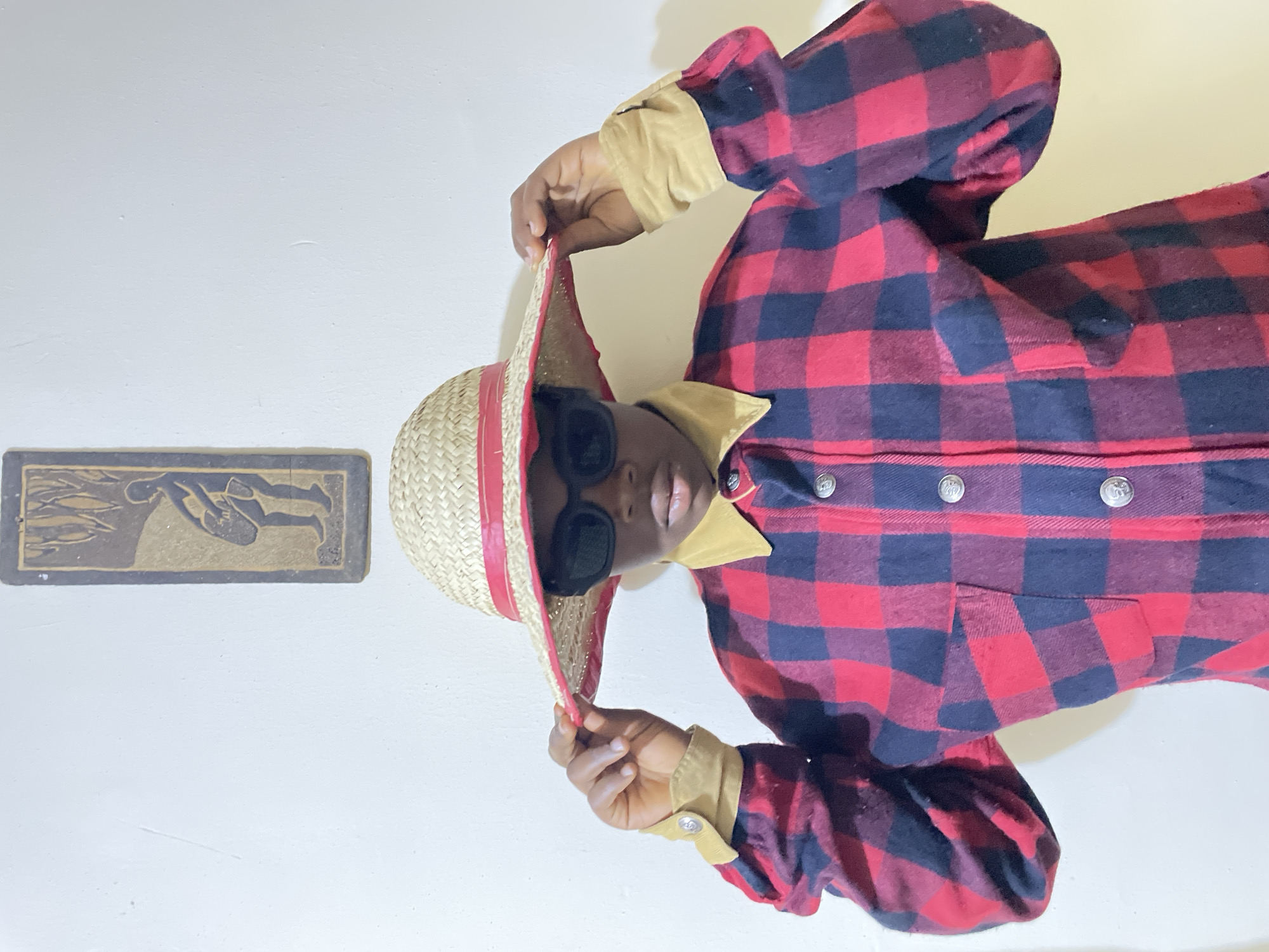 An indoor photograph, rotated 90 degrees counter-clockwise, captures a person posing against a plain, light-colored wall. The individual, who has a dark skin tone, is seen from the chest up, looking directly at the camera. They are wearing dark sunglasses and a straw hat adorned with a red band, which they grasp by the brim with their right hand.
The person is dressed in a thick, long-sleeved shirt or jacket featuring a distinctive red and dark blue (almost black) buffalo check plaid pattern. The garment has a contrasting tan or khaki-colored collar and cuffs, and several round, silver-toned buttons are visible down the front.
To the upper left of the person (when viewed in the current orientation), a rectangular art piece or plaque is mounted on the wall. This dark-toned relief sculpture depicts stylized human figures, one appearing to dive or swim alongside fish, with lighter accents highlighting the details. The background is an unadorned white or off-white wall, providing a clean, minimalist setting. The pose is casual and direct, conveying a sense of confidence. The context for this image is Jos, Nigeria.