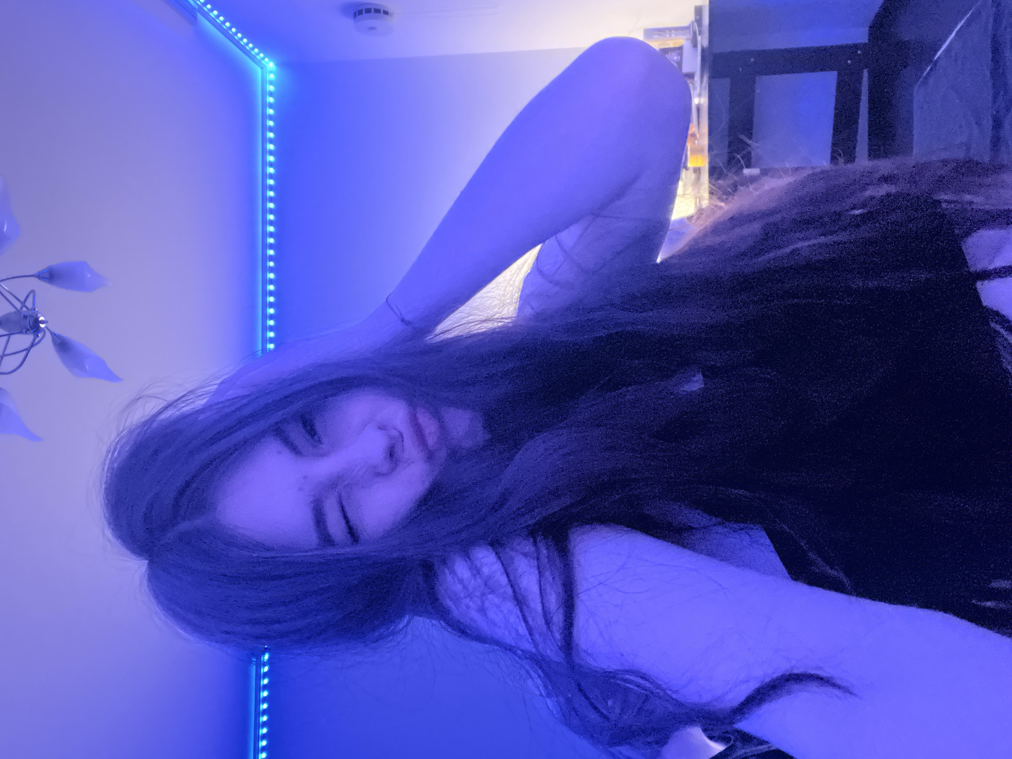 A young woman with long, dark hair is lying down, with one arm raised behind her head and the other bent by her face. Her expression is relaxed, and she is looking towards the camera with one eye slightly closed. The room is illuminated with a strong blue light from a strip of LED lights running along the wall. A ceiling fan is visible in the upper left corner, and a cabinet or shelf with objects is present in the background. The location is not discernible from the image; the city of Saint Petersburg, Russia cannot be determined.
