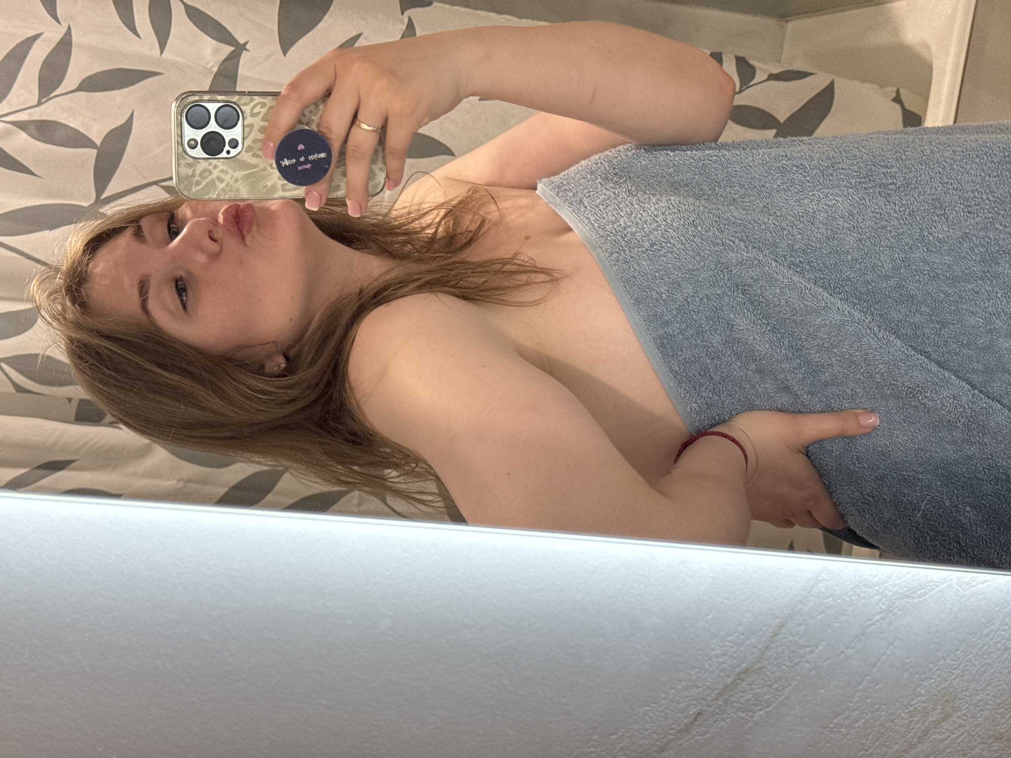 A light-skinned woman is posing in a mirror selfie. She is partially covered with a blue towel. She is looking toward the camera with puckered lips. She is wearing a gold ring on her finger and holding a phone with a pop socket. The background features a wall with a grey and white leaf pattern and the edge of a shower stall. The location is potentially a bathroom based on the towel and shower stall. The location in the city of Ферма, Russia, cannot be verified from the image.