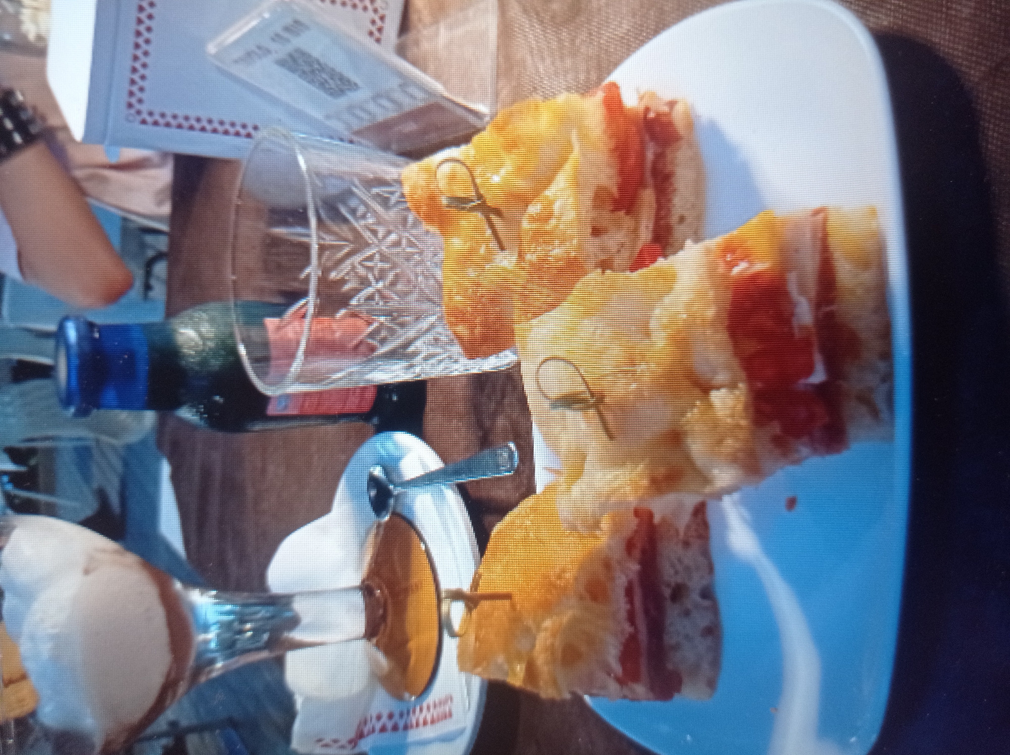 The image depicts a close-up of a white plate on a brown, textured surface. On the plate are three small, layered food items, appearing to be sandwiches or appetizers. Each item consists of bread topped with a red filling, possibly tomato or cured meat, and a lighter filling, possibly cheese or cream. Toothpicks with decorative ends are inserted into each item. In the background, a glass with a patterned texture is visible, containing a light-colored beverage. To the left of the glass, a dark blue bottle with a metallic cap is present. Further in the background, a tall glass with a dessert, possibly ice cream with chocolate sauce, is seen. A portion of a person's arm, wearing a bracelet, is visible in the upper right of the image. A white rectangular object with a red border and a QR code on it is also present in the background. The setting appears to be a cafe or restaurant. The image quality is blurry and pixelated. There is no information in the image to indicate a location in Mukachevo, Ukraine.