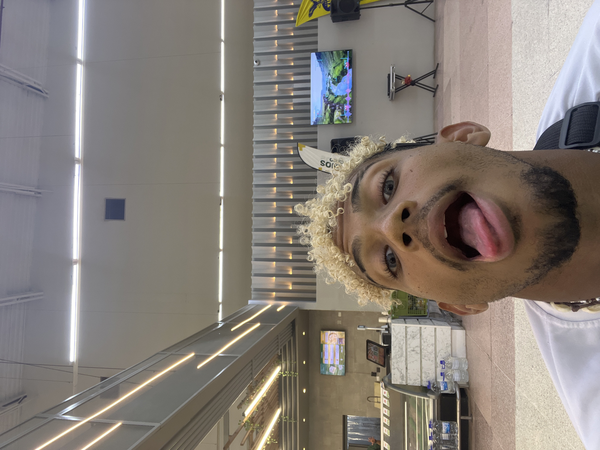 In Porlamar, Venezuela, a young man with blond, curly hair and light blue eyes is taking a selfie in what appears to be a supermarket. He is sticking out his tongue and making a funny face. Behind him, there are shelves stocked with water bottles, a refrigerated display case, and a large screen displaying a scenic landscape. The ceiling features modern lighting, and there are decorative vertical lights on the wall. The floor is tiled with a pattern of pink and beige squares.