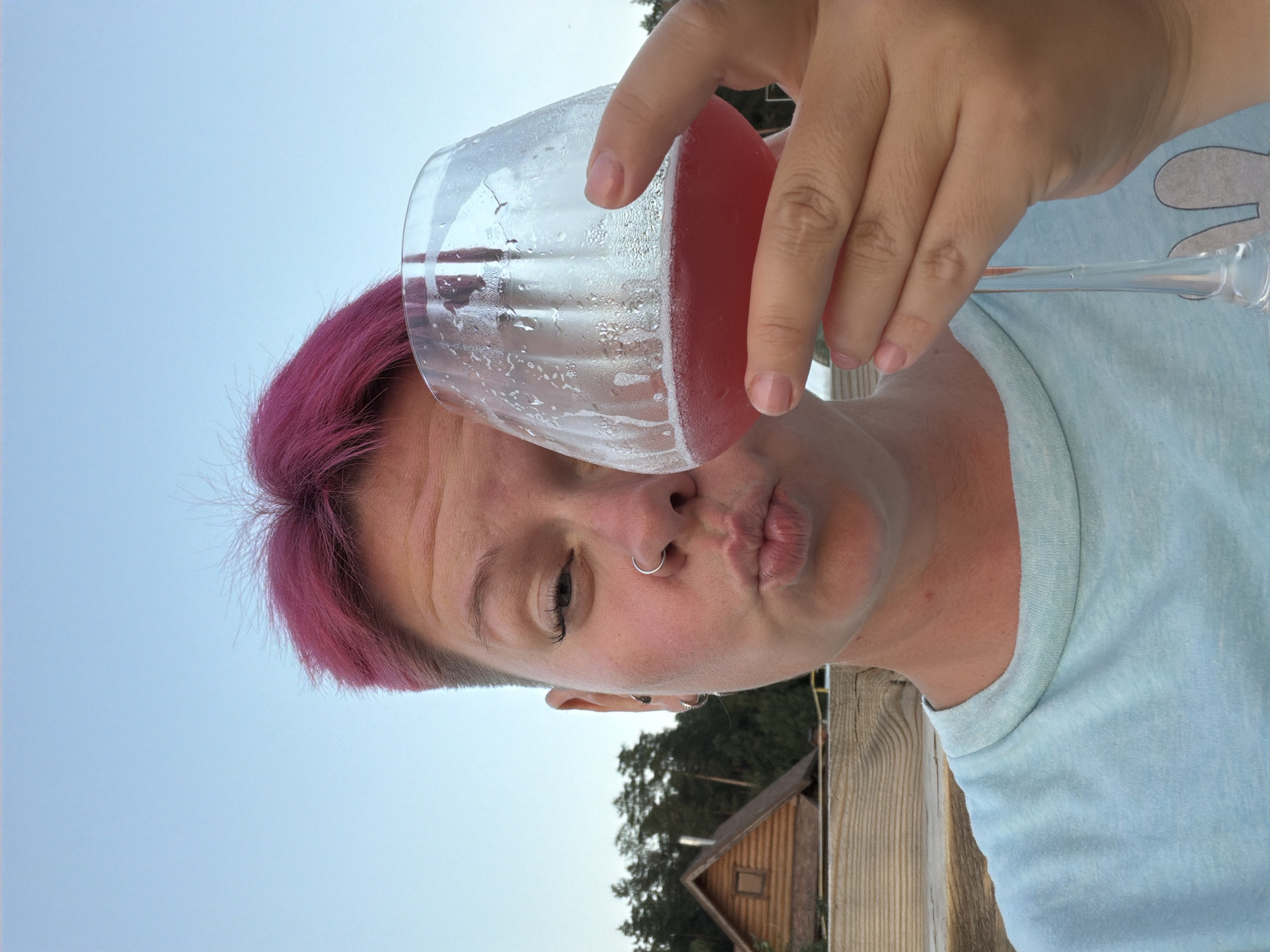 The image is a medium shot of a person with bright pink, short hair and a nose ring, holding a glass of pink liquid and making a kissing face towards the camera. The person is wearing a light blue t-shirt. In the background, there is a wooden structure resembling a small house or cabin, surrounded by trees. The sky is a clear, light blue. The image appears to have been taken outdoors, possibly in a rural or suburban setting. There are no discernible landmarks that specifically place the image in Chaikovsky, Russia, other than the initial prompt.