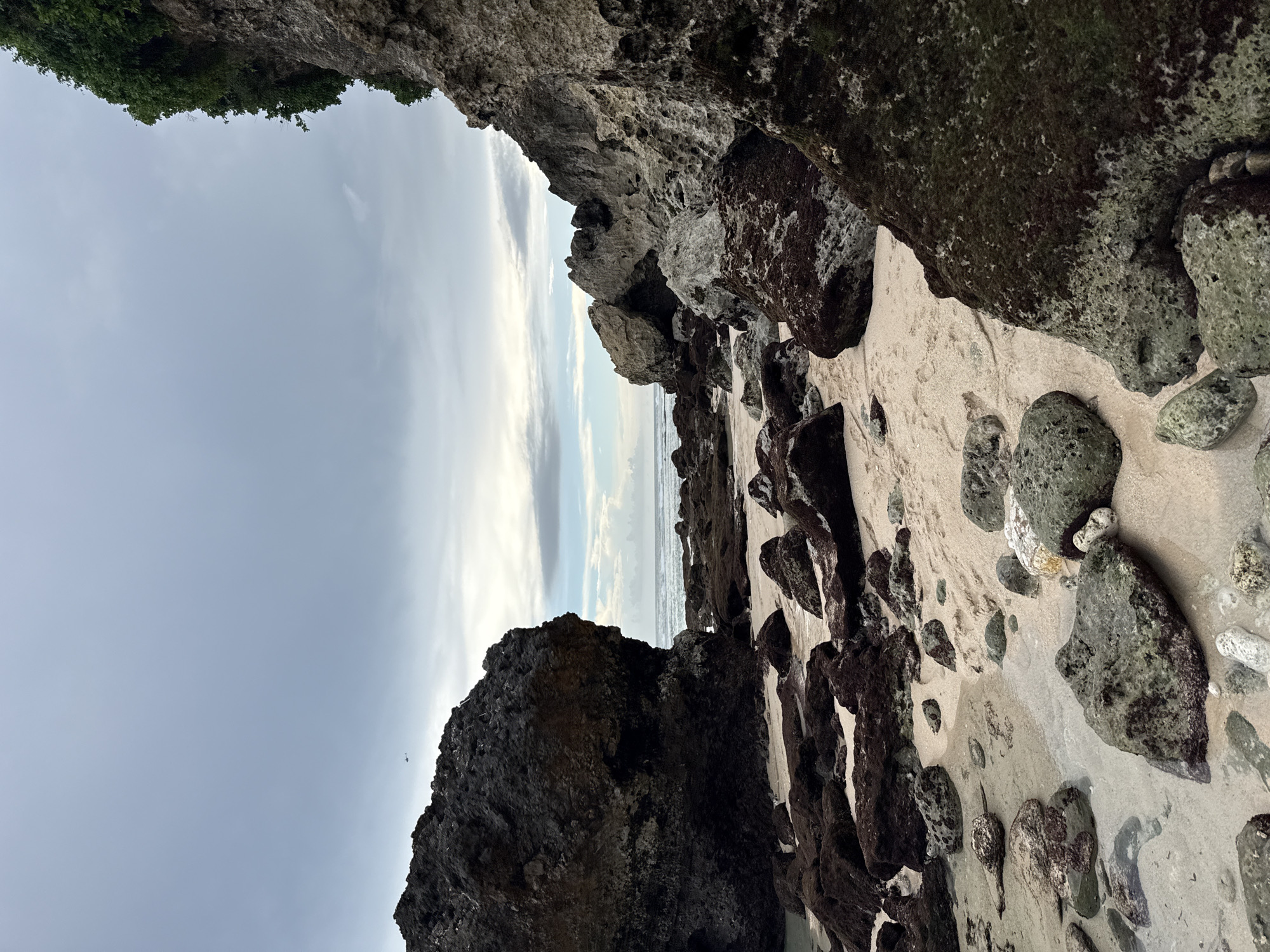 The image depicts a rocky beach in Ungasan, Indonesia. Large boulders frame the scene, with some displaying moss growth. Sand is visible between the rocks, leading toward the ocean. The sky is overcast, and a hint of vegetation is seen at the top left of the frame.