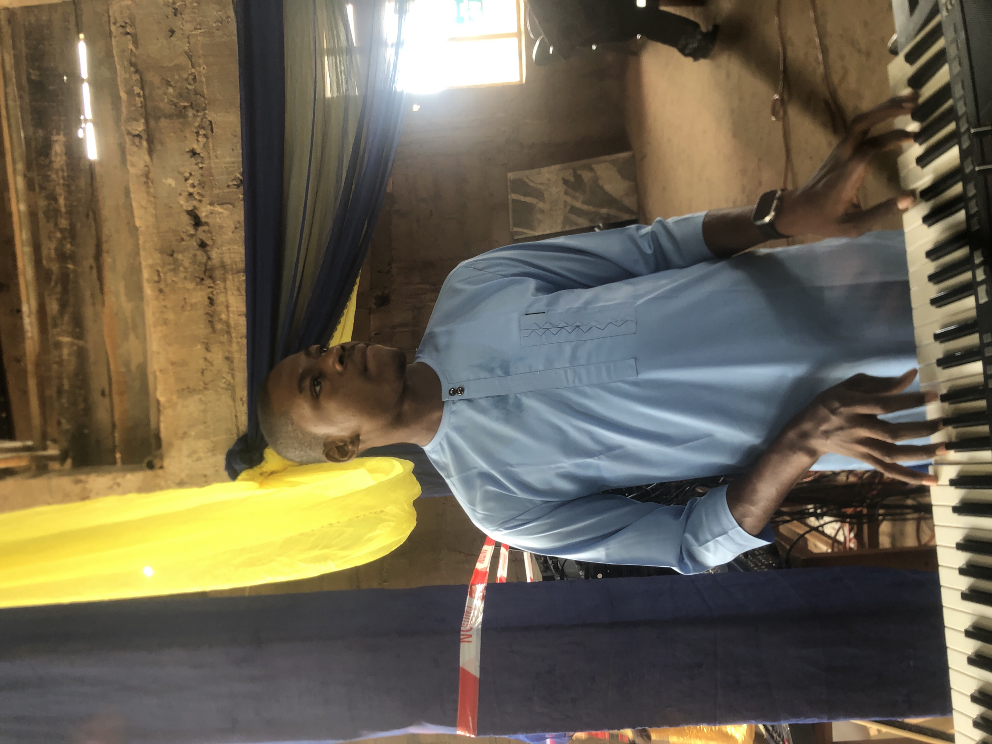 A man in a light blue shirt plays a keyboard in a room in Nteje, Nigeria. The room has rough, textured walls and is decorated with yellow and blue drapes. There is a window providing natural light. A roll of red and white caution tape is visible near the keyboard.