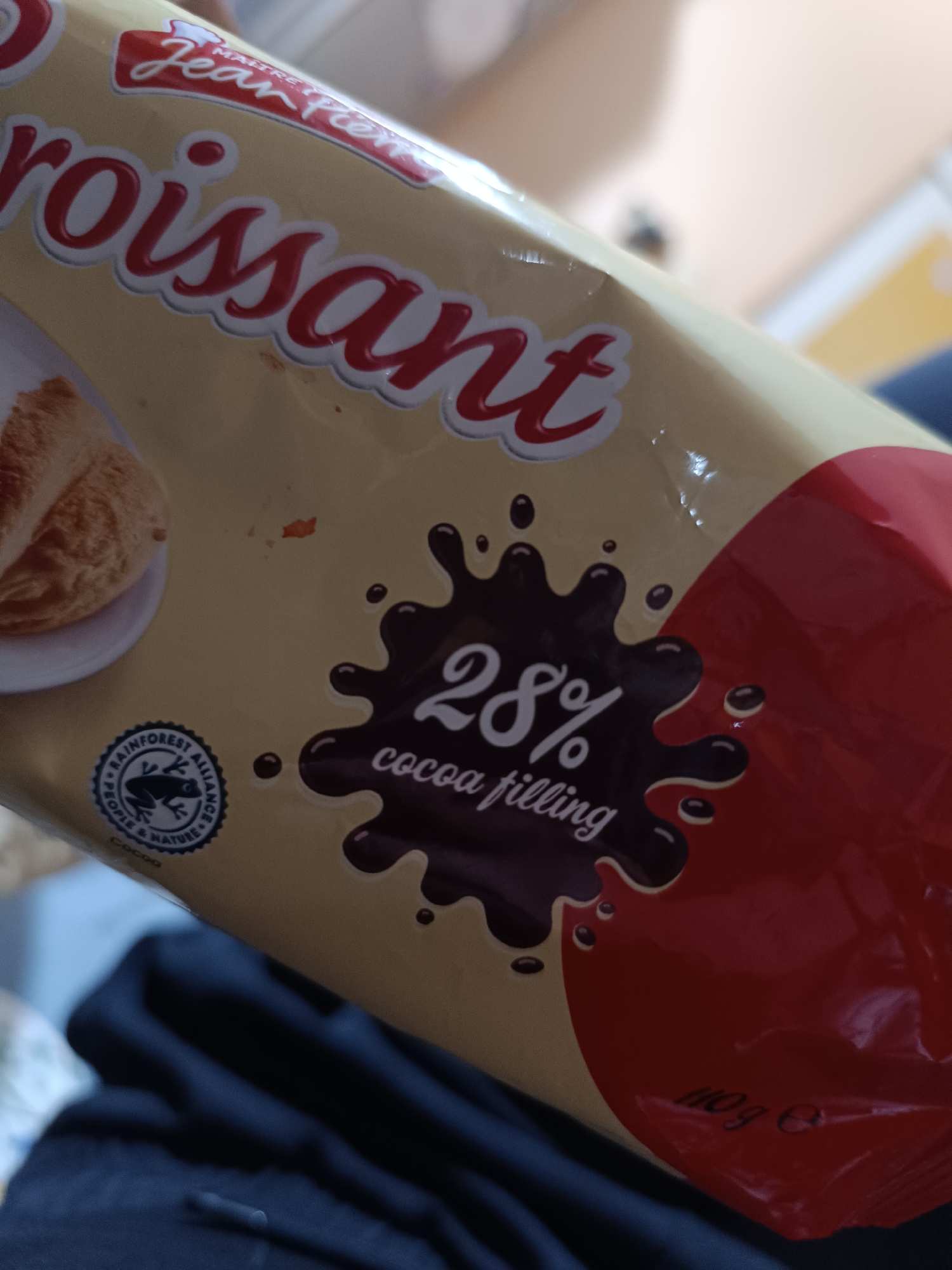 The image shows the packaging of a "Jean Pier" croissant with a cocoa filling. The package indicates that the filling contains 28% cocoa. There is a "Rainforest Alliance" logo. The croissant is red in color. It shows a chocolate blot image. The croissant is 110g. There is no city, or Bulgarian location mentioned.