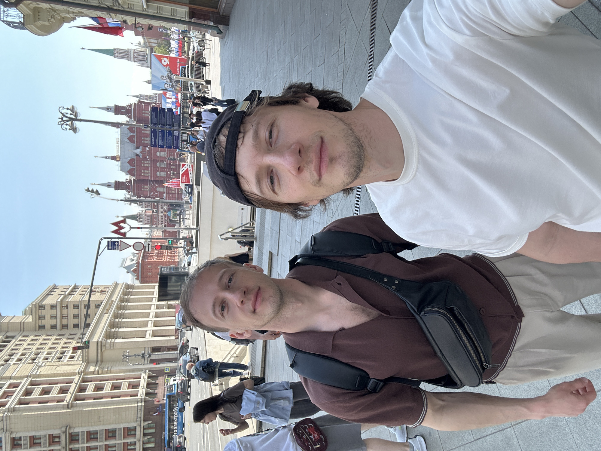 Two young men are posing for a selfie outdoors. The man on the right is wearing a white T-shirt and a dark baseball cap. The man on the left is wearing a brown, short-sleeved collared shirt, a black fanny pack, and a backpack. The Red Square in Moscow, Russia, is visible in the background. Several people are walking in the background. The day appears to be sunny.