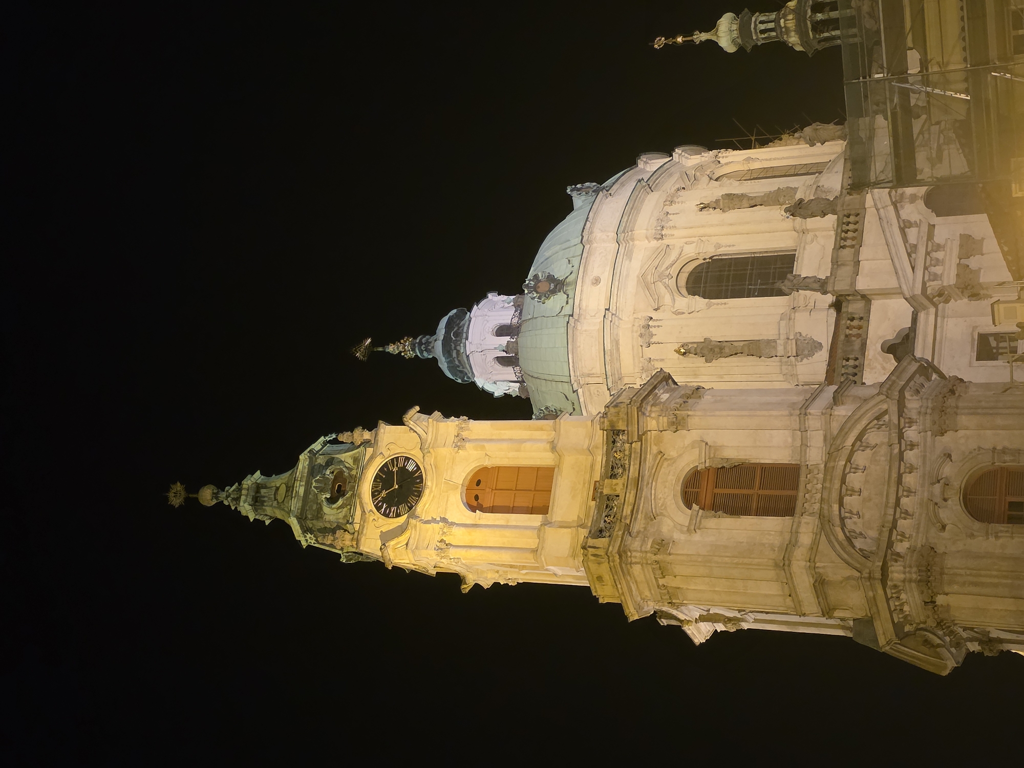 Captured at night, the image showcases the illuminated architectural grandeur of St. Nicholas Church in Prague. The camera, positioned at an upward angle, accentuates the height and ornate detailing of the Baroque structure against a stark black sky. The clock tower prominently displays a functioning clock face, while the church's dome rises elegantly behind it. Artificial lights cast a warm glow, highlighting the intricate facades, windows, and sculptural elements, underscoring the historic and aesthetic significance of this landmark.