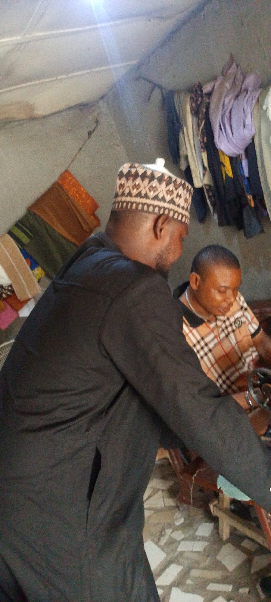 In a room in Lokoja, Nigeria, two men are present. One man, wearing a black robe and a patterned kufi cap, stands with his back to the camera. The other man, wearing a plaid shirt, is seated at a sewing machine. Clothes hang from a makeshift clothesline in the background. Other pieces of fabric are draped nearby. The room has a concrete ceiling and tiled floor.
