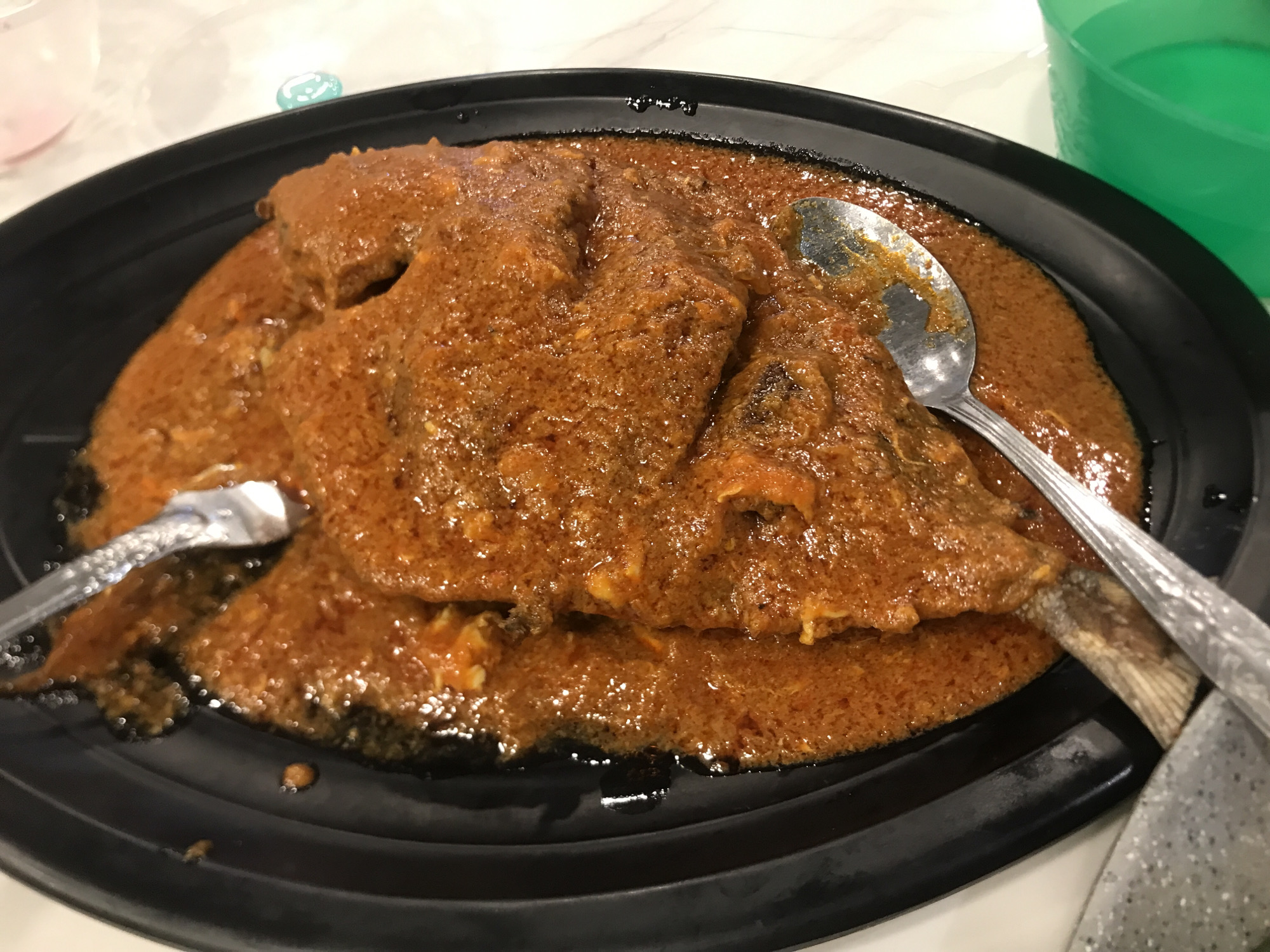 A plate of Asam Pedas fish dish on a black serving platter with a spoon and fork. The scene is Blangbitra, Indonesia. The fish is covered in a thick, reddish-brown sauce. The metal spoon and fork are partially submerged in the sauce. A green container is visible in the background.