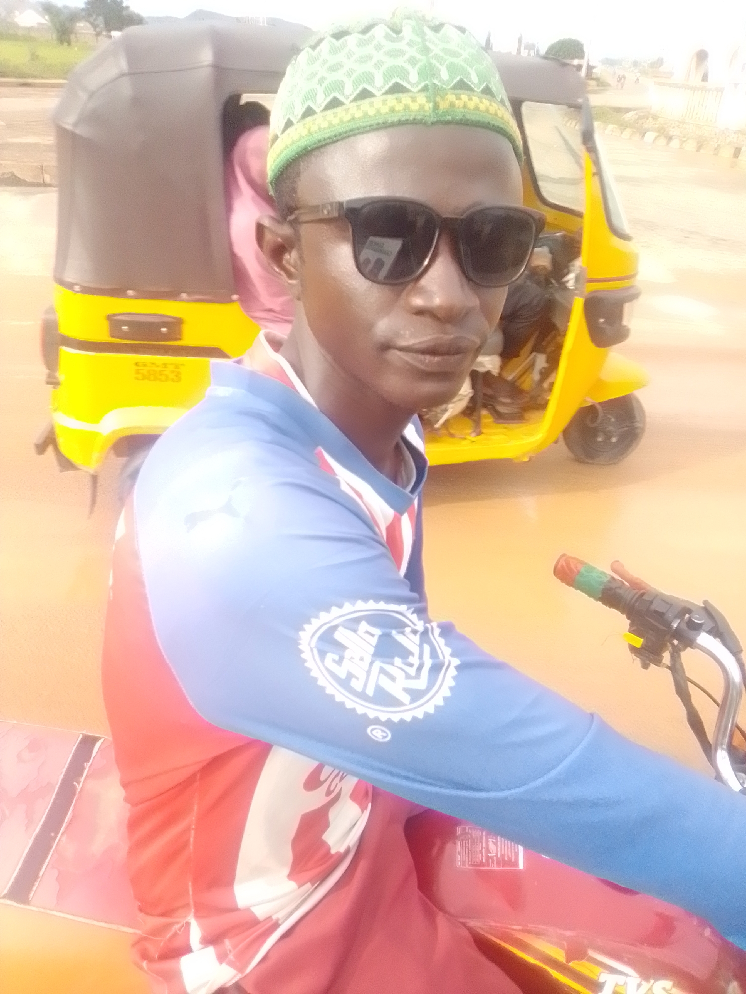 In Kwadon, Nigeria, a young man wearing a green and yellow patterned kufi hat and dark sunglasses is pictured. He is seated on a red and yellow motorcycle. He is wearing a blue long-sleeved shirt with white and red accents on the chest and arm. A yellow and black auto-rickshaw is parked behind him. The ground is paved with asphalt, and there is a slight haze in the air.