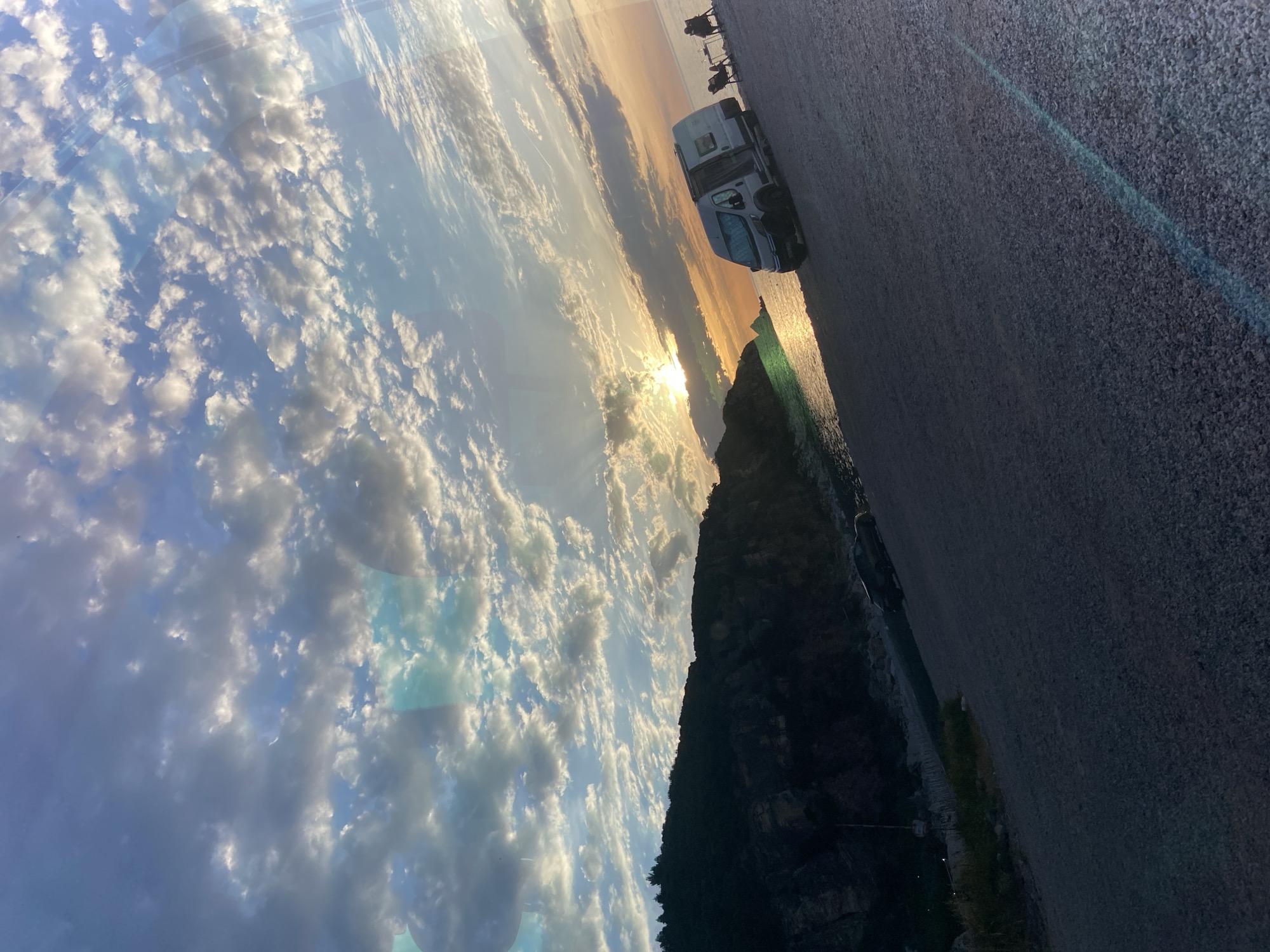 The image depicts a coastal scene at either sunrise or sunset, heavily tilted counter-clockwise. A low sun is positioned near the horizon on the right, casting a bright golden reflection path across the calm water. The sky is largely overcast with various cumulus and stratocumulus clouds, transitioning from blue and grey in the upper-left to orange and golden hues near the sun.
On the left, dark, steep hills or mountains covered with sparse vegetation and visible rock strata form the coastline, silhouetted against the sky. A dark-colored passenger vehicle is partially visible parked on a road or track at the base of these hills.
In the midground, a light-colored camper van is parked on a flat, gravelly or rocky surface directly adjacent to the water on the right side. The van's side door is open, and a sunshade is visible on its windshield. To the right of the camper van, two individuals are seated in camping chairs next to a small table, facing the water and the setting/rising sun. Their figures are mostly silhouetted.
The immediate foreground consists of the dark, textured gravel surface. A distinct blue lens flare is present in the bottom right corner, extending diagonally across the frame.