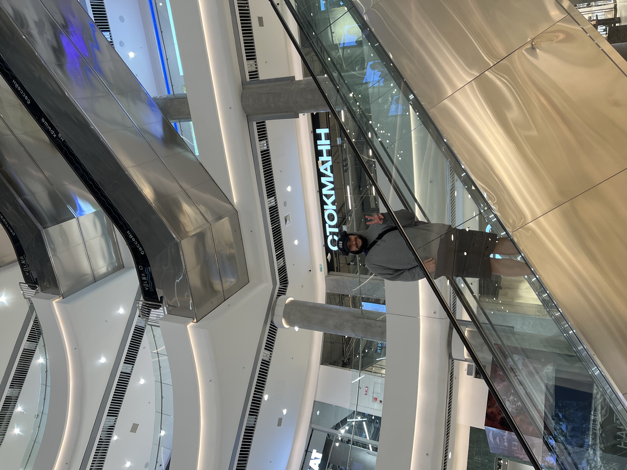 This image captures a man's reflection in the glass railing of an escalator within a brightly lit, modern multi-story shopping mall. The man, likely taking a selfie, is wearing a gray hoodie, dark shorts or pants, and a dark beanie or cap, and is making a peace sign with his right hand.
The setting is characterized by contemporary architecture with prevalent use of white walls, shiny metallic escalator surfaces, and glass railings. Bright artificial lighting from various fixtures illuminates the expansive interior. Large, curved structural elements and pillars are visible throughout the space.
In the background, a prominent black sign with white text reads "СТОКМАНН" (STOCKMANN), indicating a department store. Other reversed text is visible on the sides of the escalators due to reflections. These include "БАССЕЙН" (SWIMMING POOL), "VOLNA" (WAVE), and "РЕСТОРАН" (RESTAURANT), further confirming the commercial nature of the building as a shopping complex. The atmosphere appears to be active and bustling, typical of a mall during its operating hours, although no other people are explicitly visible besides the man's reflection. The image is taken in Khabarovsk, Russia.