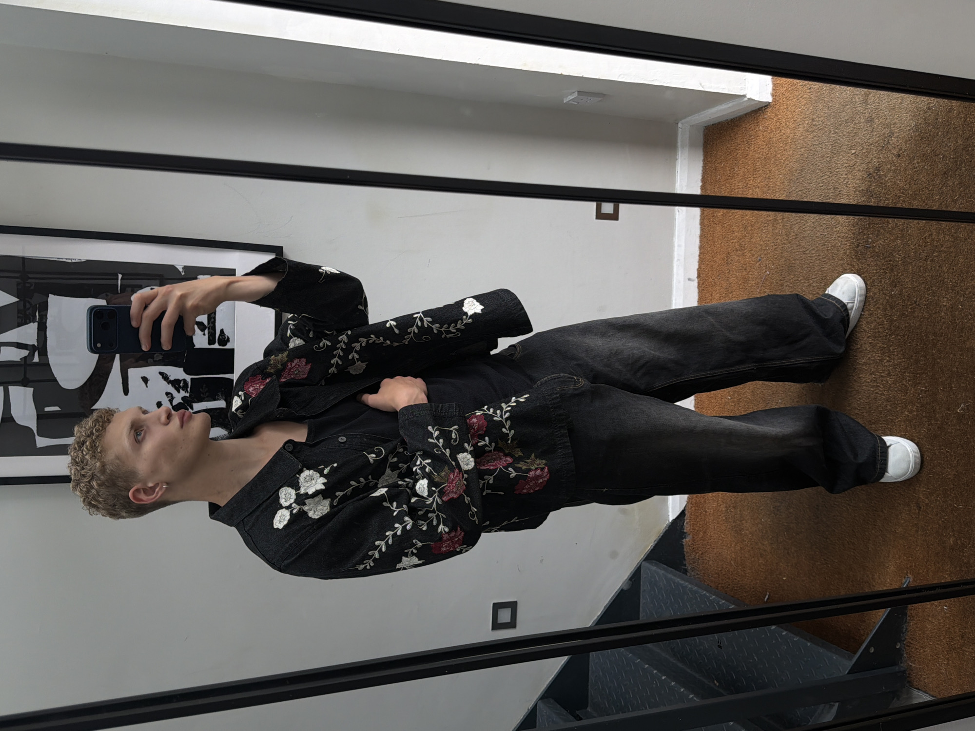A young individual, appearing male, with short curly blonde hair and light-colored eyes, is depicted in a full-length mirror selfie. They wear a dark denim jacket extensively embroidered with white, red, and green floral patterns, layered over a plain black undershirt. This is paired with loose-fitting, dark grey denim jeans and white low-top sneakers. A small silver hoop earring is visible in the left ear. The individual holds a dark-colored smartphone in their right hand, angled towards the mirror, indicative of taking the photograph. Their left hand is tucked into the front of their jacket, and their gaze is directed towards the right side of the frame.
The environment, reflected in the mirror, features plain light grey walls. A large, framed black and white abstract artwork hangs on the wall behind the individual's head. To the left, a dark, industrial-style metal staircase with diamond plate treads ascends upwards. The floor is covered with a textured, burnt orange carpet. A dark square light switch or outlet is mounted on the wall adjacent to the staircase. On the right side of the reflection, a white Type G electrical outlet is visible near the floor, above a white baseboard. The presence of a Type G electrical outlet indicates the scene is located in the United Kingdom, specifically in an urban setting such as London.