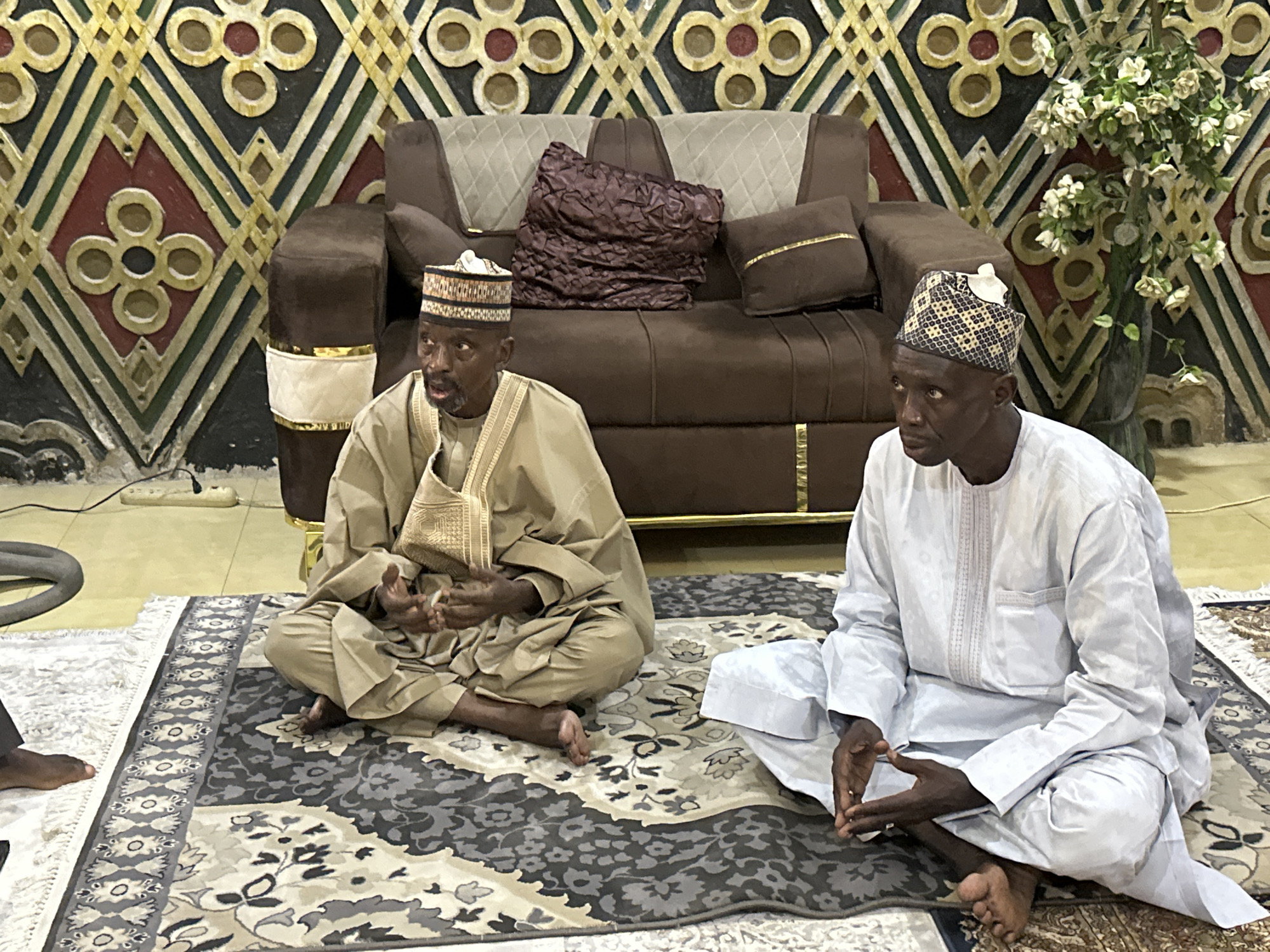 Two men are seated cross-legged on a large, patterned rug indoors. The man on the left is wearing a beige traditional outfit with a matching hat and has a small pouch or bag hanging from his shoulder. The man on the right is dressed in a white outfit with a patterned hat. Behind them is a brown sofa with decorative pillows. The wall features an intricate geometric design. A vase with white flowers is visible in the background to the right. A vacuum cleaner is partially visible on the left side of the rug. A black foot is partially visible on the left side of the rug. The city and exact location are unknown.