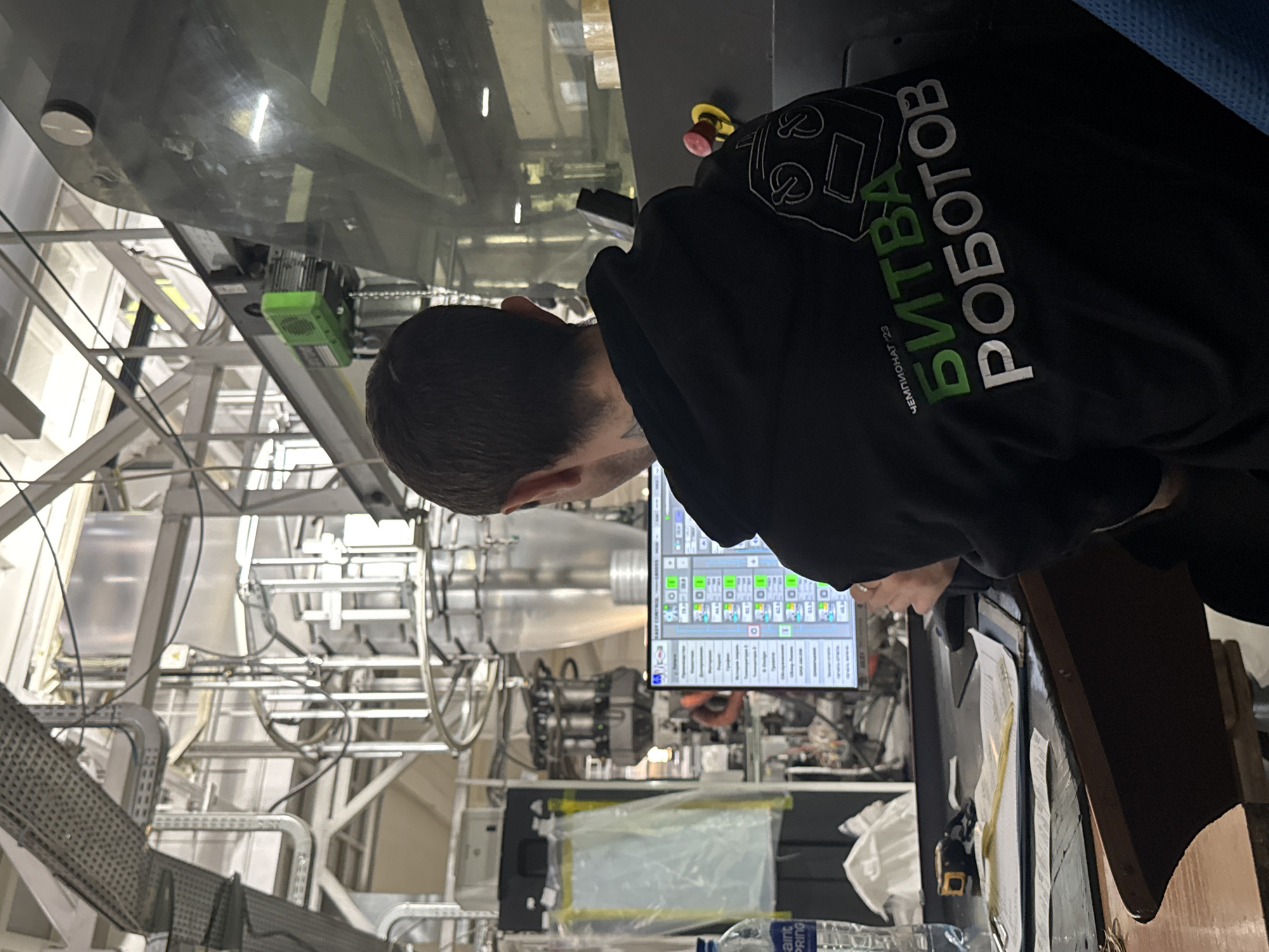 A person is standing in front of a large piece of machinery, possibly in an industrial setting. The machine features a control panel with a digital display that shows several icons and parameters. The machinery itself appears complex, with tubes, wires, and mechanical parts. The person is wearing a black hoodie with the Russian text "Битва Роботов" (Battle of Robots) and "Чемпионат 23" (Championship 23), along with a logo of a robot, suggesting the image was taken at or related to the Battle of Robots Championship in Perm, Russia. A water bottle and papers are visible on a surface in the foreground.