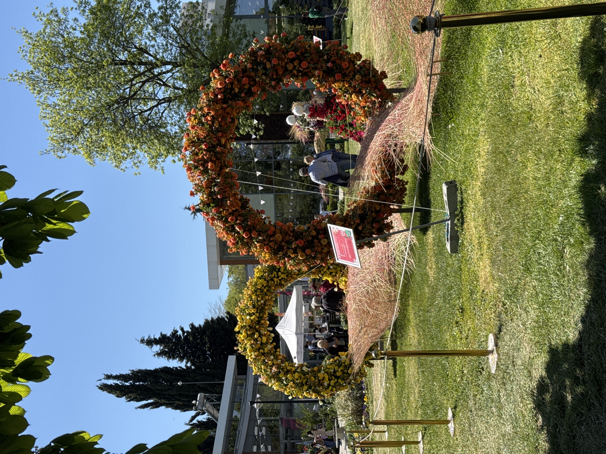 The image shows two floral arches on a grassy area, likely in a park or garden, with people walking around in the background. The top arch is decorated with orange flowers, and the lower arch is decorated with yellow flowers. The arches are supported by golden poles and have a backdrop of dried grass-like material. A sign is visible next to the orange flower arch. The location is in the city of Burgas, Bulgaria. Trees and a clear blue sky are visible in the background.