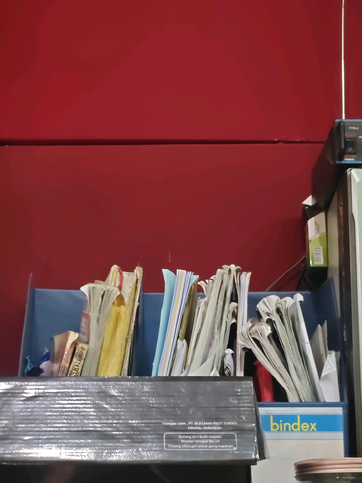 The image depicts a shelf with various documents and files stored in a blue organizer. A black box is placed in front of the organizer, with text indicating it is from PT. Bersama Pasti Sukses, Jakarta, Indonesia. A small electronic device with an antenna and power button is visible on the right side of the shelf, next to a light green box. The background is a solid red wall. The scene appears to be an indoor setting, possibly an office or storage area. No people or actions are visible. The location is stated to be in Sumberwono, Indonesia, but this is not visually confirmed within the image itself.