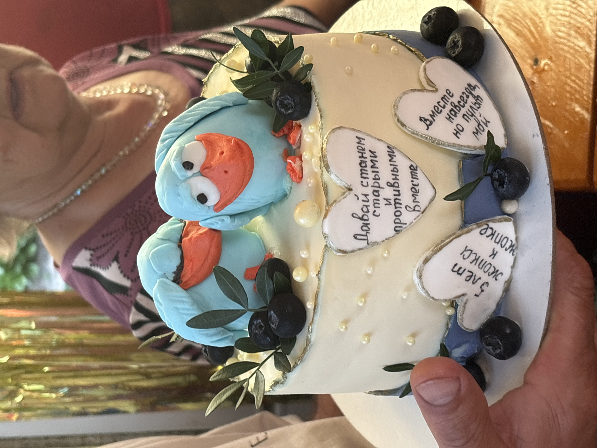 In Rozhdestveno, Russia, a round, two-tiered cake with white and light blue frosting is presented from a low-angle perspective. The cake is adorned with two light blue, cartoonish bird figures with orange beaks and white eyes on top. The cake's sides are decorated with small green leaf sprigs, dark blueberries, and scattered white and gold edible pearls. Three heart-shaped plaques with Cyrillic text are affixed to the side: the top heart reads "Let's become old and obnoxious together," the bottom left reads "5 years ass to ass," and the bottom right reads "Together forever, but the remote is mine." A light-skinned hand, likely male, supports the cake from below. In the blurred background, an elderly female with light hair, wearing a purple patterned top and a beaded necklace, is partially visible. Golden metallic streamers and subtle hints of natural light and greenery are present in the far background. A wooden surface is partially visible in the lower right foreground.