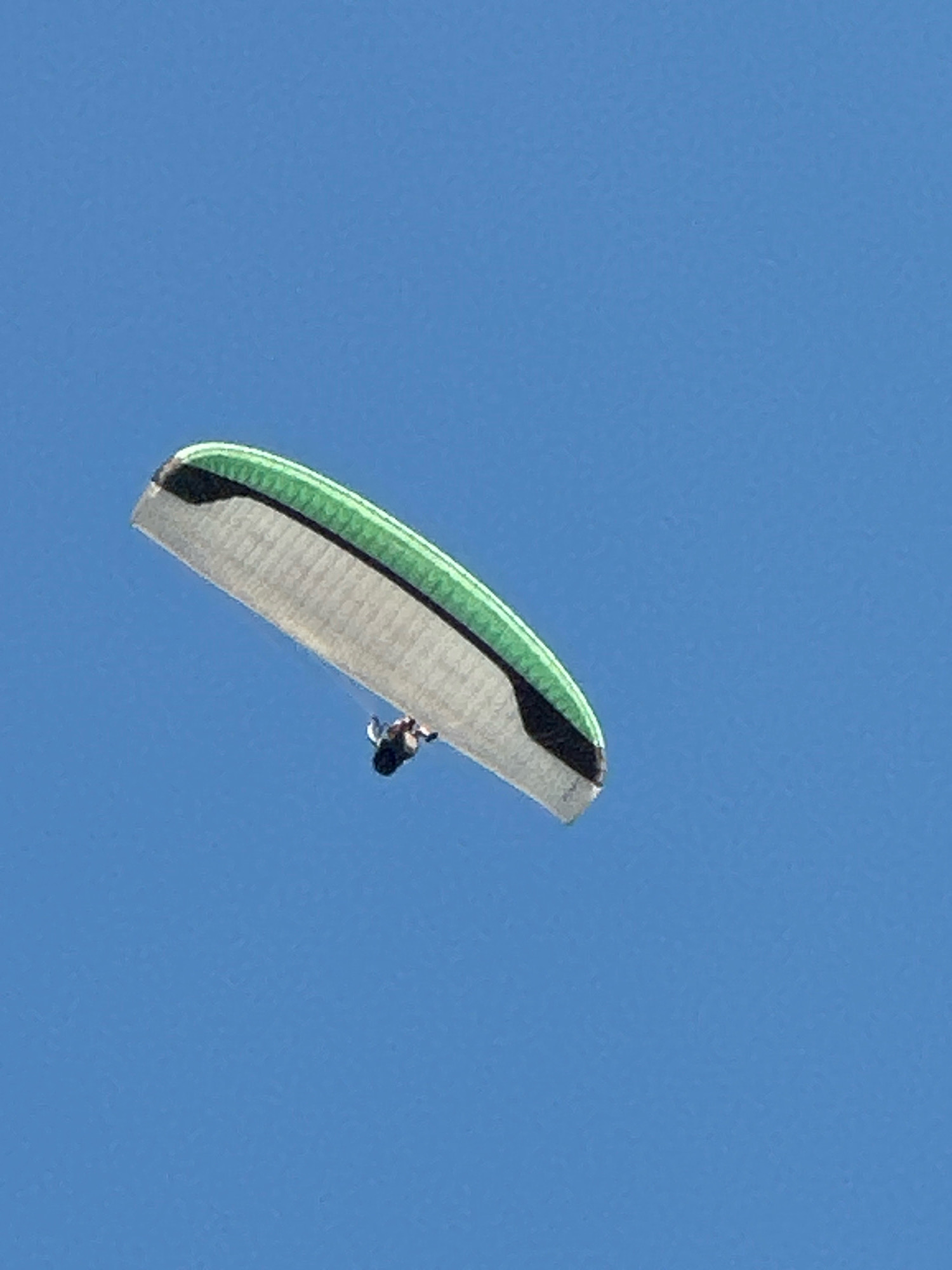 A paraglider is captured in mid-air against a clear blue sky. The paraglider, a person suspended in a harness beneath a colorful wing, appears to be descending or maneuvering. The wing of the paraglider is predominantly a light gray, with a vibrant green stripe along its leading edge and a dark band on the trailing edge. The individual in the harness is small and indistinct due to the distance. There are no discernible landmarks or environmental features to indicate a specific location within the city of Boreti, Montenegro, or any other city. The image focuses solely on the paraglider in its aerial environment.