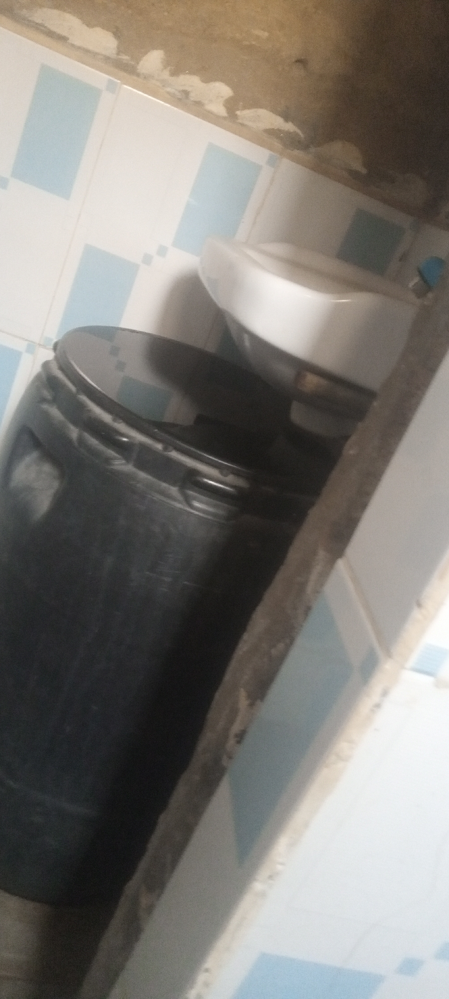 The image shows a bathroom interior. A black plastic drum with a lid is visible, positioned close to a white ceramic sink mounted on a wall with blue and white tiles. The room appears cramped. The location is unspecified.