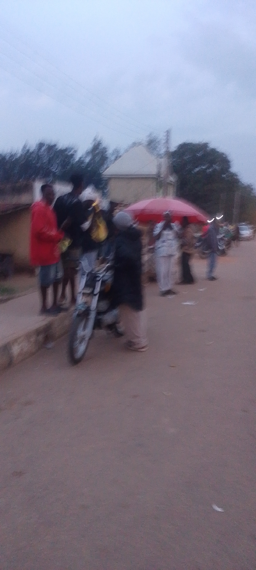 The image depicts a street scene with several people and a motorcycle. The setting appears to be outdoors in a city, likely Gura Top, Nigeria, based on the provided context.
In the foreground, a motorcycle is partially visible, with its front wheel and handlebars distinct. Behind the motorcycle, a group of individuals are gathered. One person on the left is wearing a red jacket and dark shorts, standing near a sidewalk. To their right, a person in a black shirt and light-colored shorts is also present. Further back, more people are visible, some under a bright red umbrella. One individual under the umbrella is wearing a white and patterned shirt with light-colored pants. Another person is wearing a dark outfit, possibly a robe or dress. The ground is a paved surface, likely a road, with some debris scattered on it. The background shows buildings and trees, with a cloudy or overcast sky. The overall lighting suggests it might be dusk or an overcast day. The image quality is somewhat blurred, making detailed facial features and precise actions difficult to discern.
