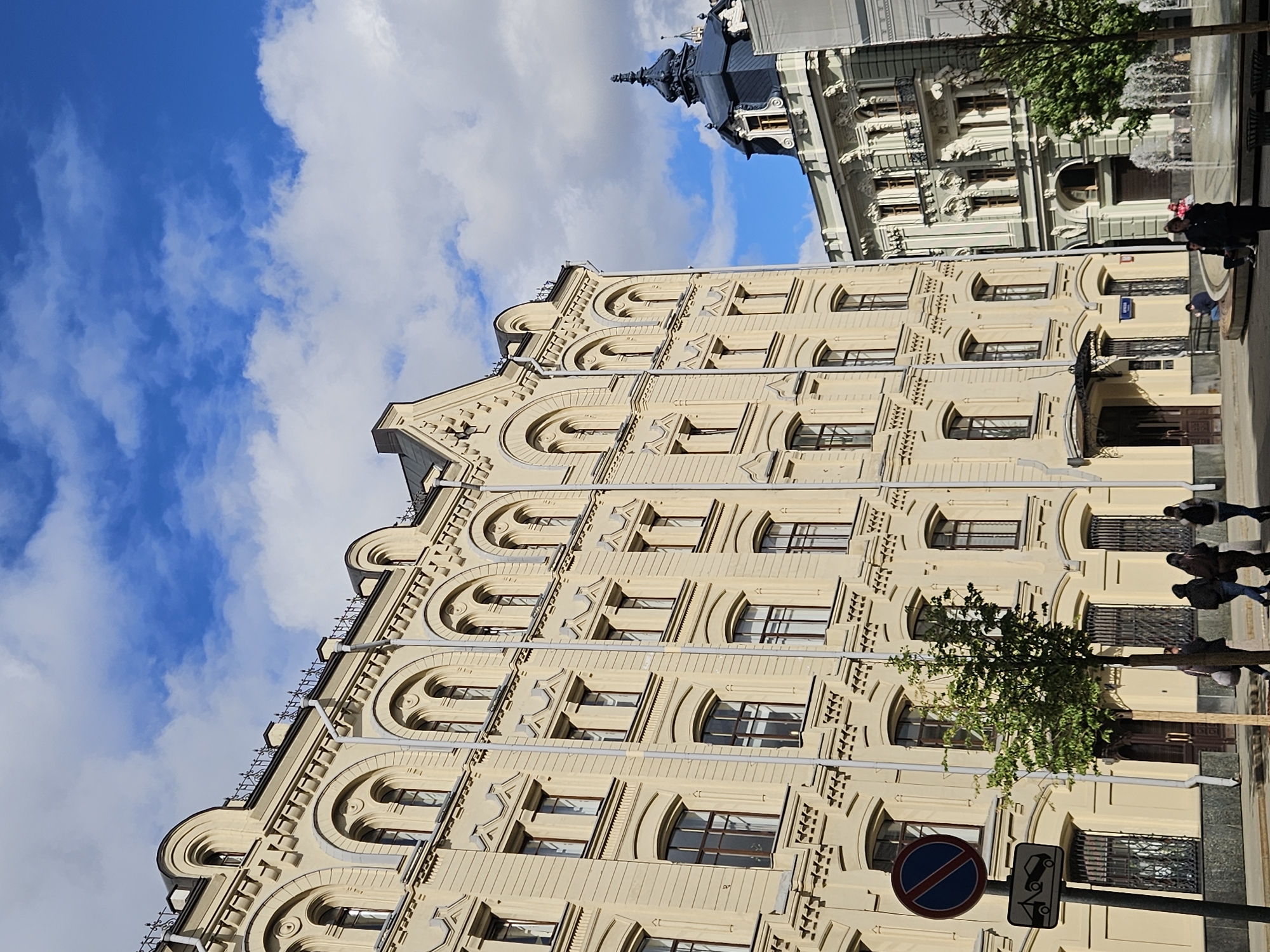 The image shows a street view in Moscow, Russia, prominently featuring a multi-story, beige building with arched windows. The sky is visible, displaying blue patches interspersed with clouds. Adjacent to the beige building is another building with a different architectural style. There are people walking along the sidewalk, and a tree is visible near the buildings. Street signs are also present.