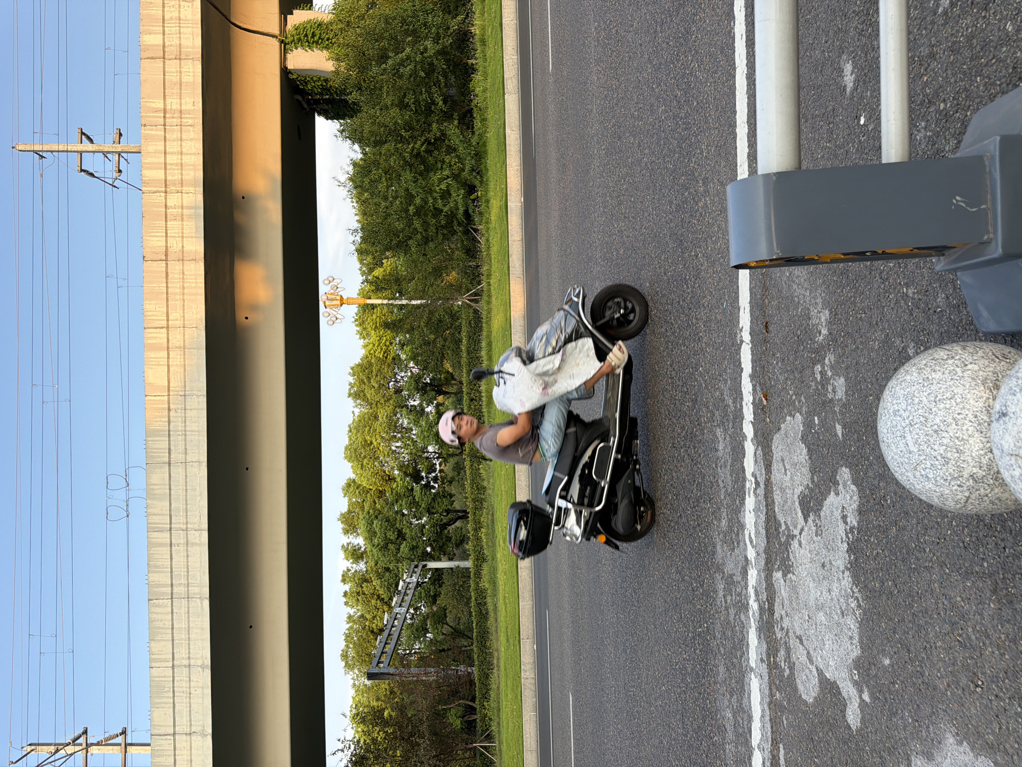 In the city of Jiangjin, China, a person wearing a pink helmet rides a black scooter down a paved road. The person is wearing a gray t-shirt and blue jeans, and is carrying a white bundle on the scooter. The scooter has a black storage box on the back. The road is bordered by a grassy median and trees. An elevated concrete structure with overhead wires runs across the background. A lamppost with multiple lights is visible behind the trees. The sky is clear and blue. The image appears to be taken during daylight hours.