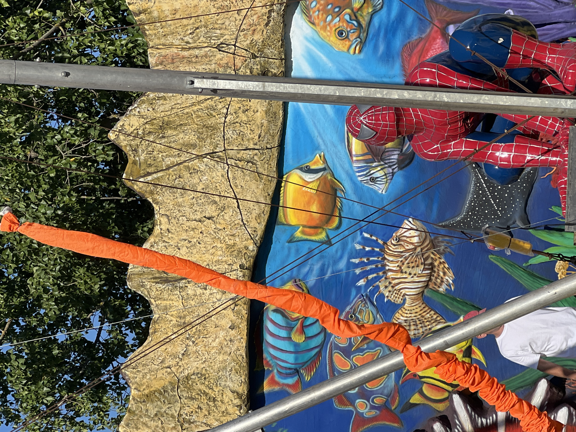 This image depicts a Spiderman figurine positioned in front of a mural of an underwater scene, specifically in Golubitskaya, Russia. The mural features various colorful fish, including angelfish, lionfish, and what appears to be a manta ray. Behind the mural, a rocky cliff formation is visible, painted in earthy tones. An orange, crinkled fabric tube is draped diagonally across the scene. Two vertical metallic poles are present, one to the left and one to the right of the Spiderman figure, and thin wires are suspended between them and attached to the mural. A person wearing a white t-shirt and a red cap is partially visible to the left of the image, behind the metallic pole. The scene is illuminated by bright sunlight, suggesting it is outdoors.