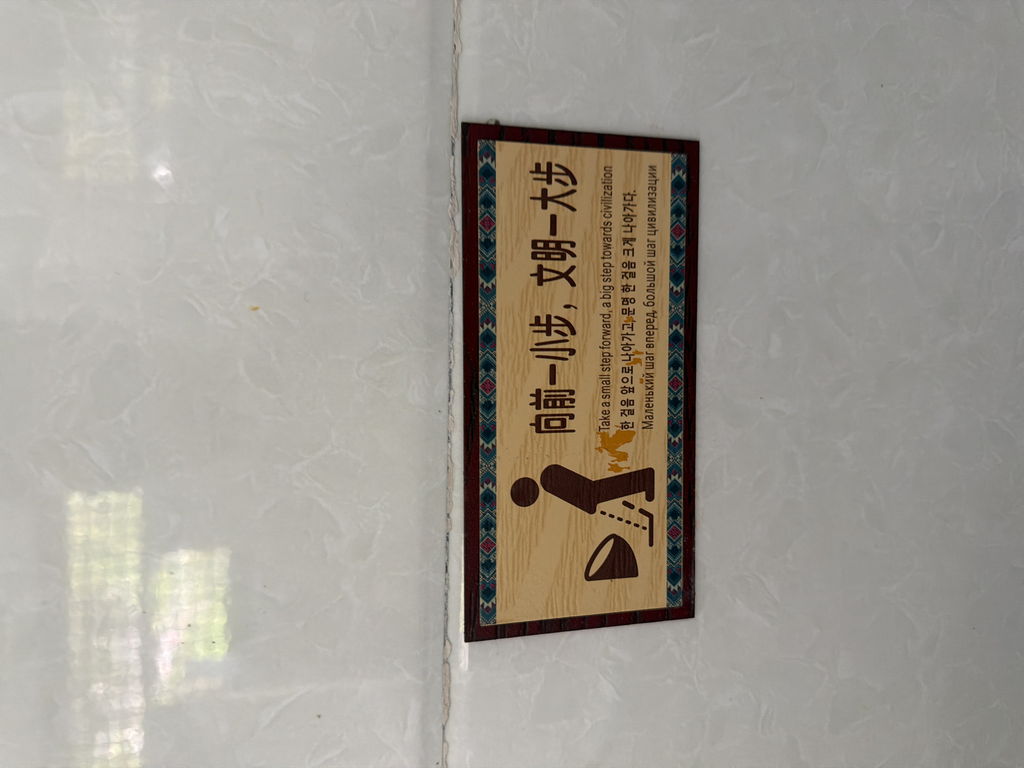 A framed sign is mounted on a light-colored textured wall. The sign itself has a wood-grain appearance with a decorative border. It includes an illustration of a figure dropping an object into a container. Above the illustration, the text reads "向前一小步,文明一大步" in Chinese, "Take a small step forward, a big step towards civilization" in English, "한 걸음 앞으로 나아가고, 문명 한 걸음 크게 나아가다." in Korean, and "Маленький шаг вперед, большой шаг цивилизации" in Russian. Based on information available, the city of Zhonghu, China is a potential location.