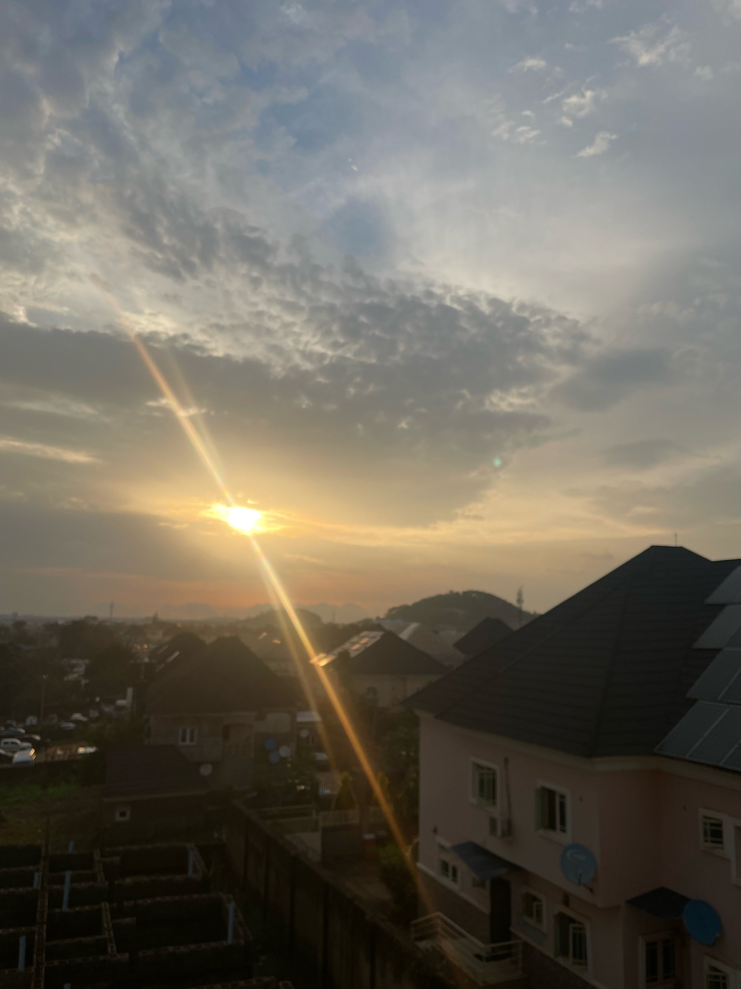 This is a photograph taken from an elevated position, likely a rooftop, in Abuja, Nigeria, during either sunrise or sunset. The sky is filled with a mix of light and dark clouds, with the sun prominently visible as a bright, glowing orb emitting distinct sunbeams that stretch across the frame.
The foreground of the image shows several residential buildings. A prominent pink building with a dark tiled roof is situated on the right side, featuring multiple windows, air conditioning units, and satellite dishes. To the left and in the mid-ground, other houses with dark roofs and various architectural features are visible, some partially obscured by trees. The lower left section of the image reveals the foundation of a building under construction, with visible concrete structures and pipes.
The overall scene depicts a suburban or semi-urban environment within Abuja, characterized by residential development and a natural landscape dominated by the dramatic sky and sunlight. The image conveys a sense of calm and the beauty of natural light.